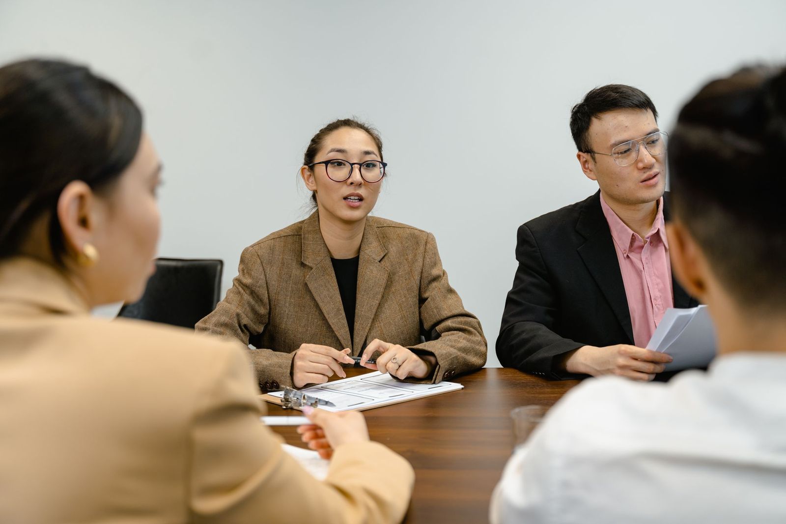 Two colleagues sitting across a table, one speaking while pointing at a printed page, others listening with papers in hand
