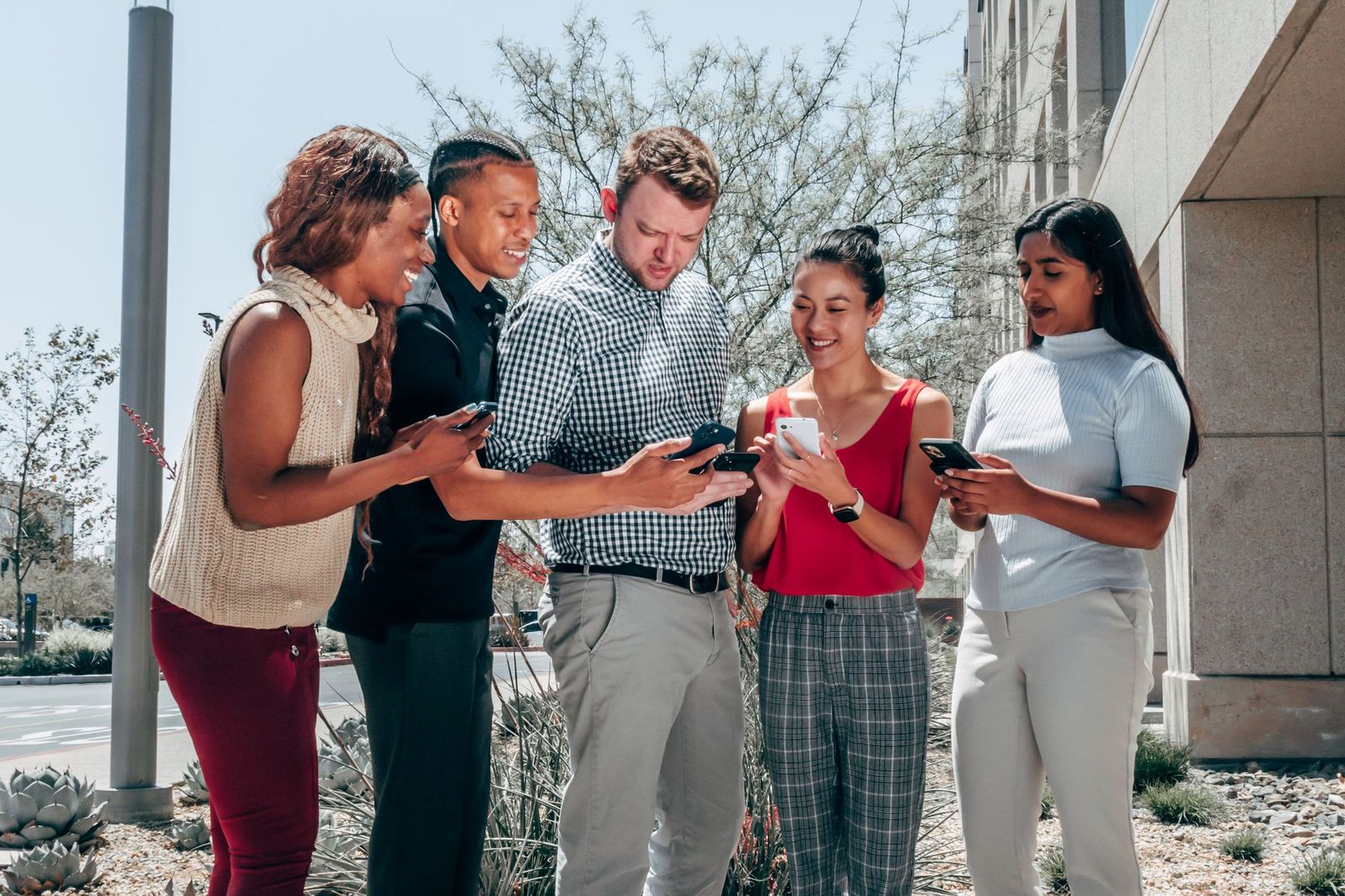 Five friends standing outdoors checking their phones together, smiling, next to a leafy street