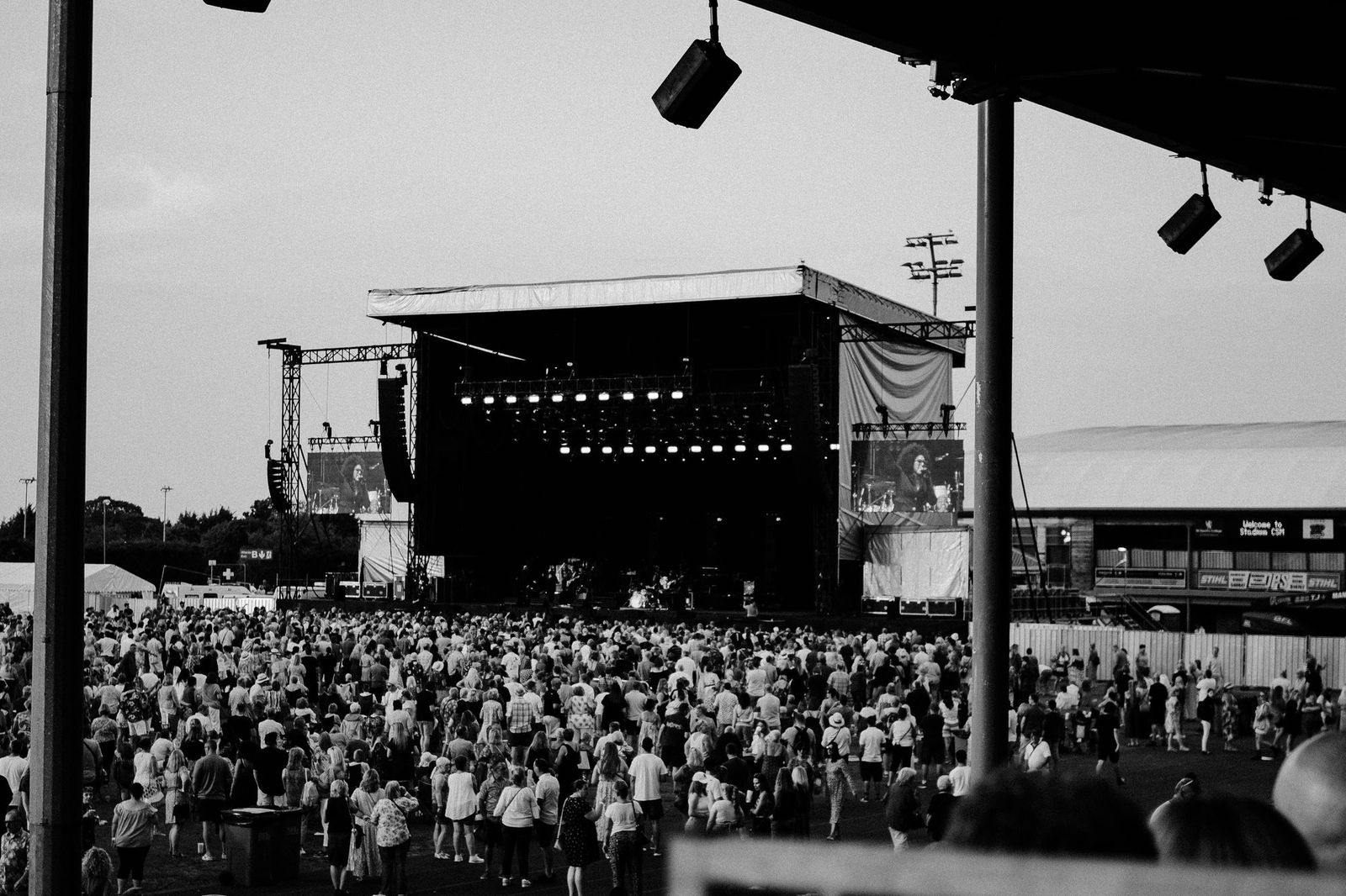 A large crowd viewed from behind facing a concert stage at dusk, in black and white