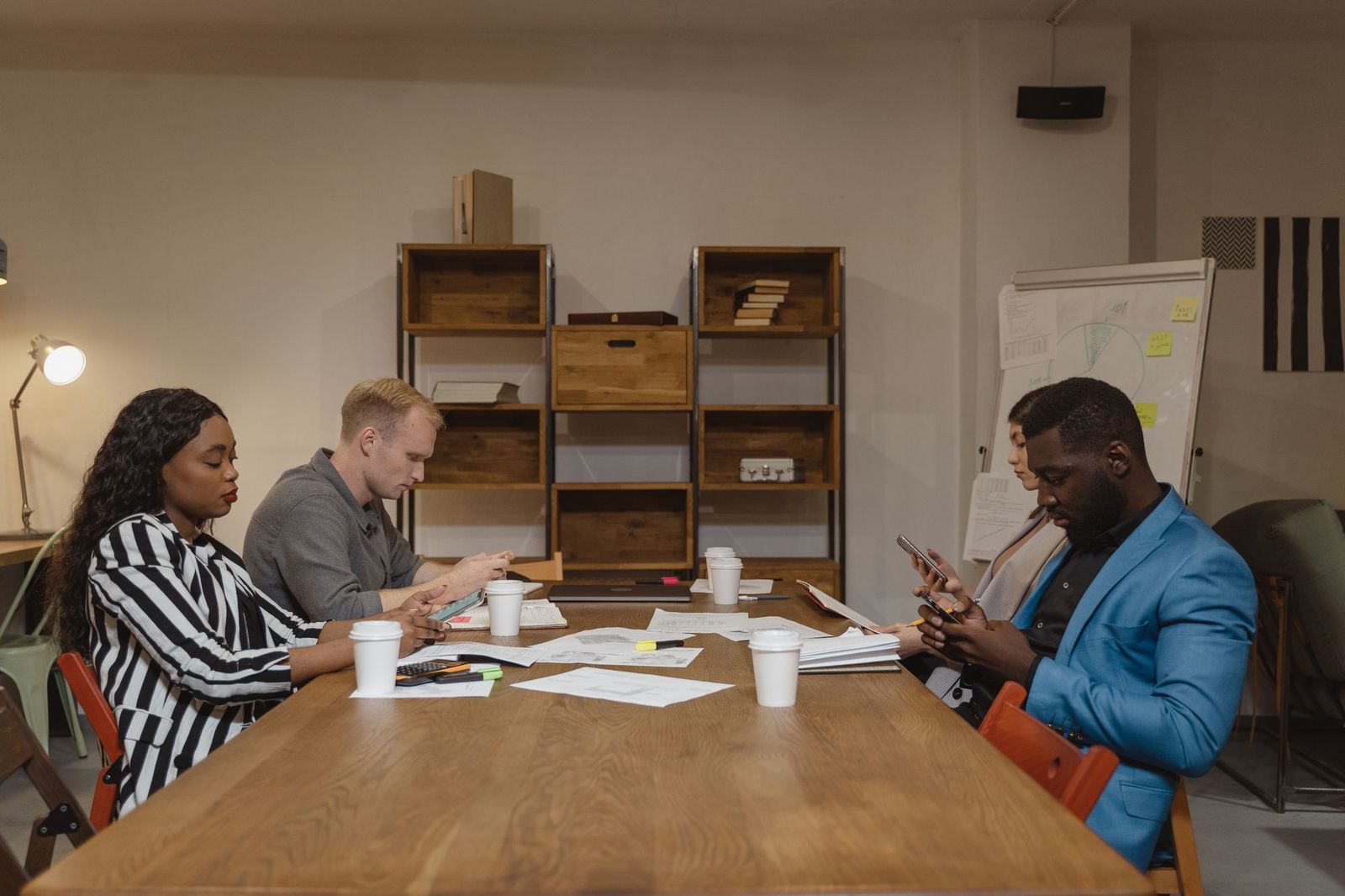 Four colleagues sitting around a table covered in papers and coffee cups, each looking down at their own phone instead of the documents in front of them