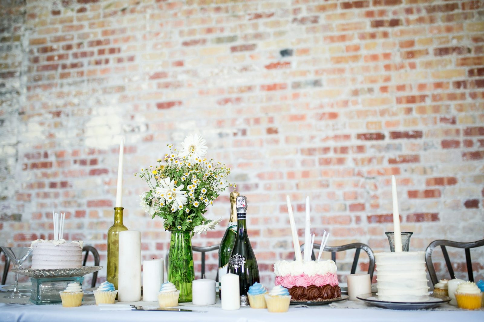 A dessert and drinks table set up against a brick wall with cakes, candles and flowers, ready before guests arrive