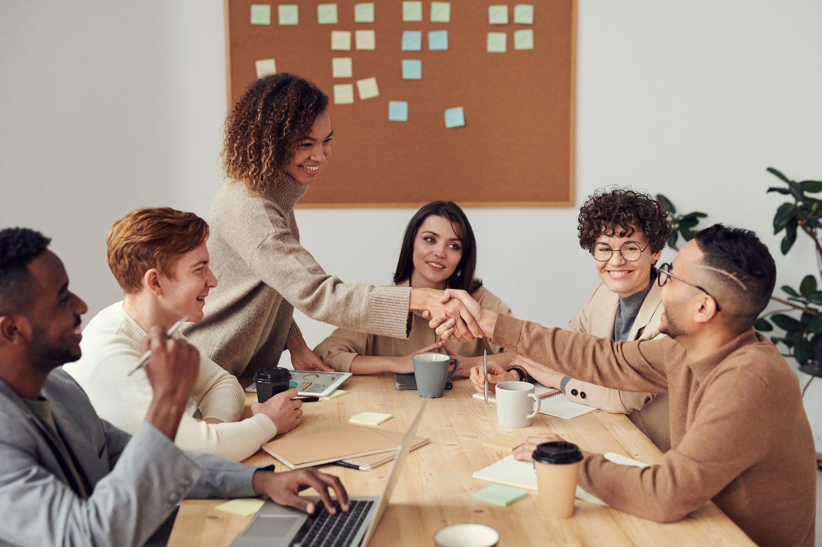 A group meeting around a table with a wall of sticky notes behind them, two people shaking hands