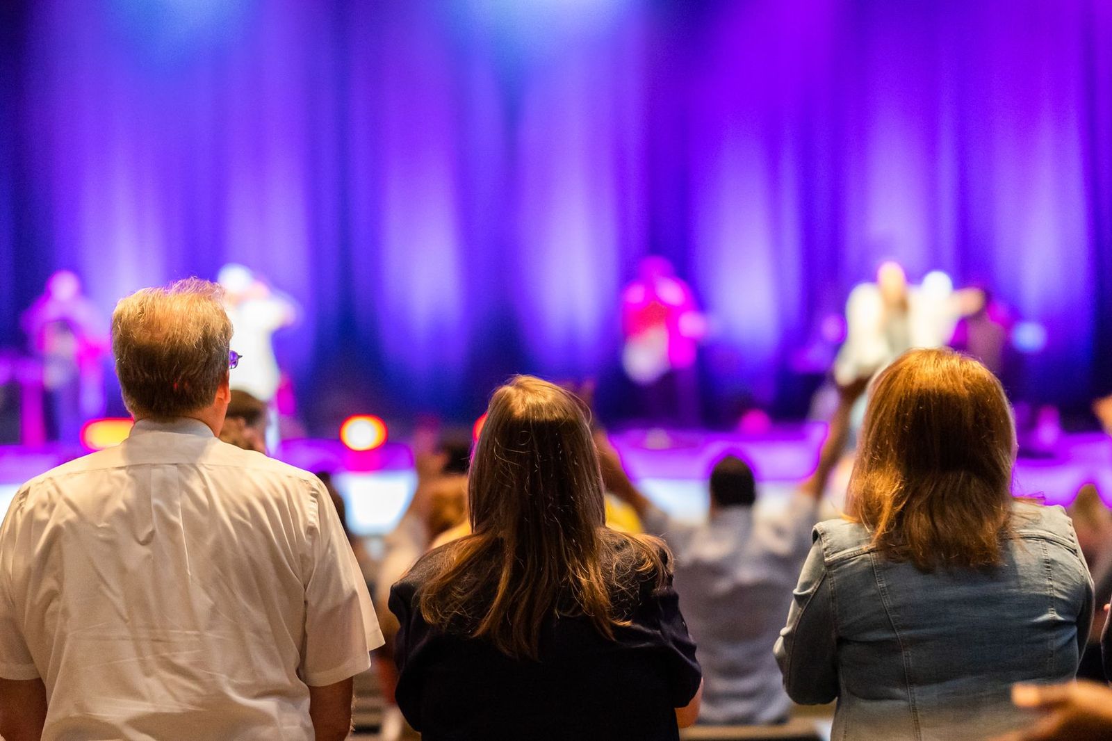 A crowd of people seen from behind, watching a lit stage from a distance in a large venue