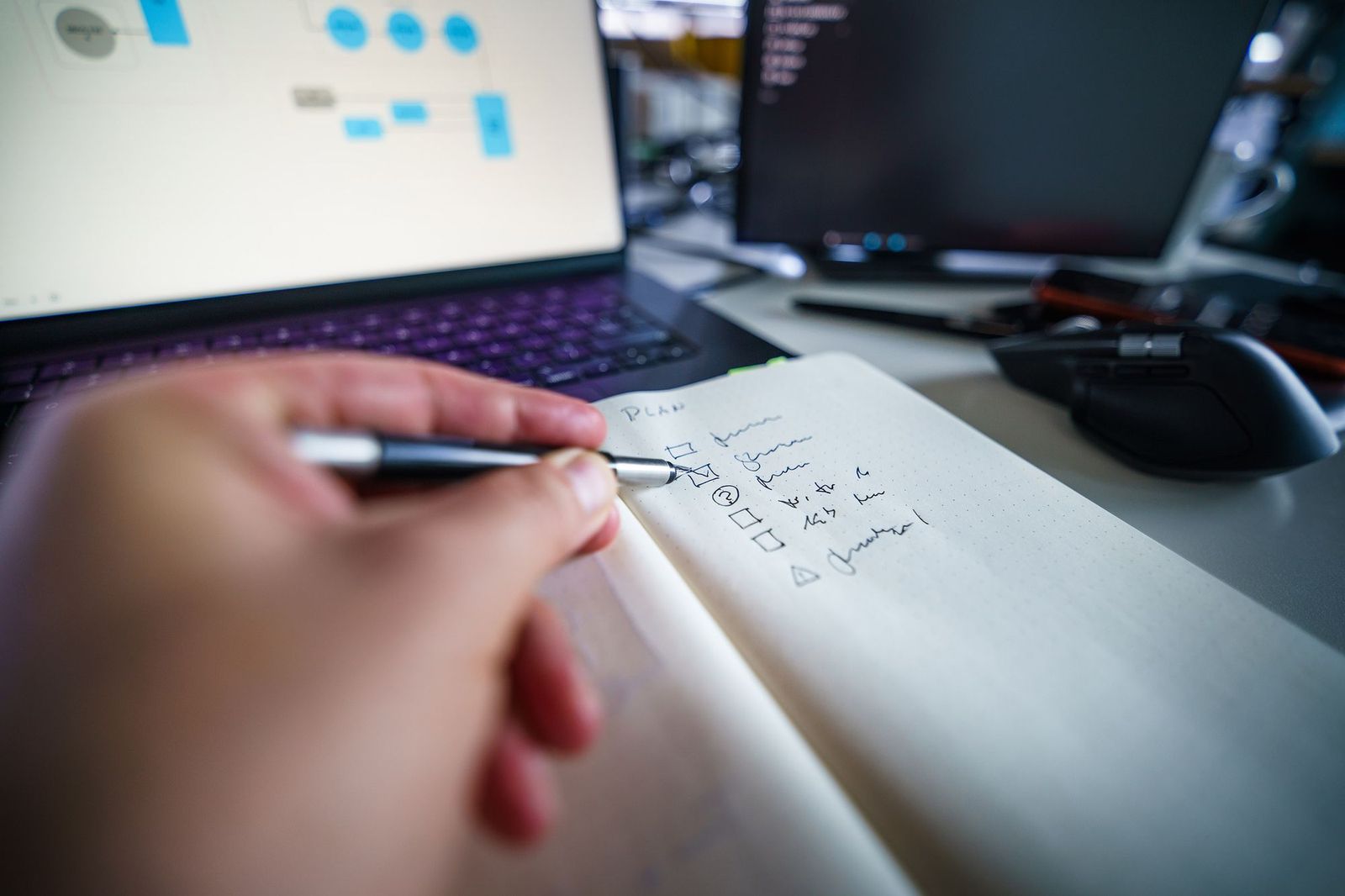 A hand writing a checklist by pen in a notebook, laptop and monitor screens with charts blurred in the background