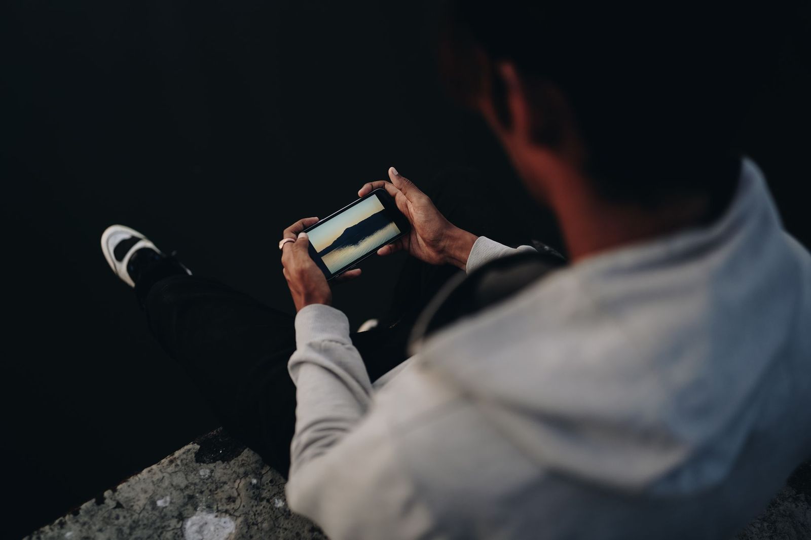 A person sitting on a stone ledge outdoors at dusk, photographing a hillside view on their phone