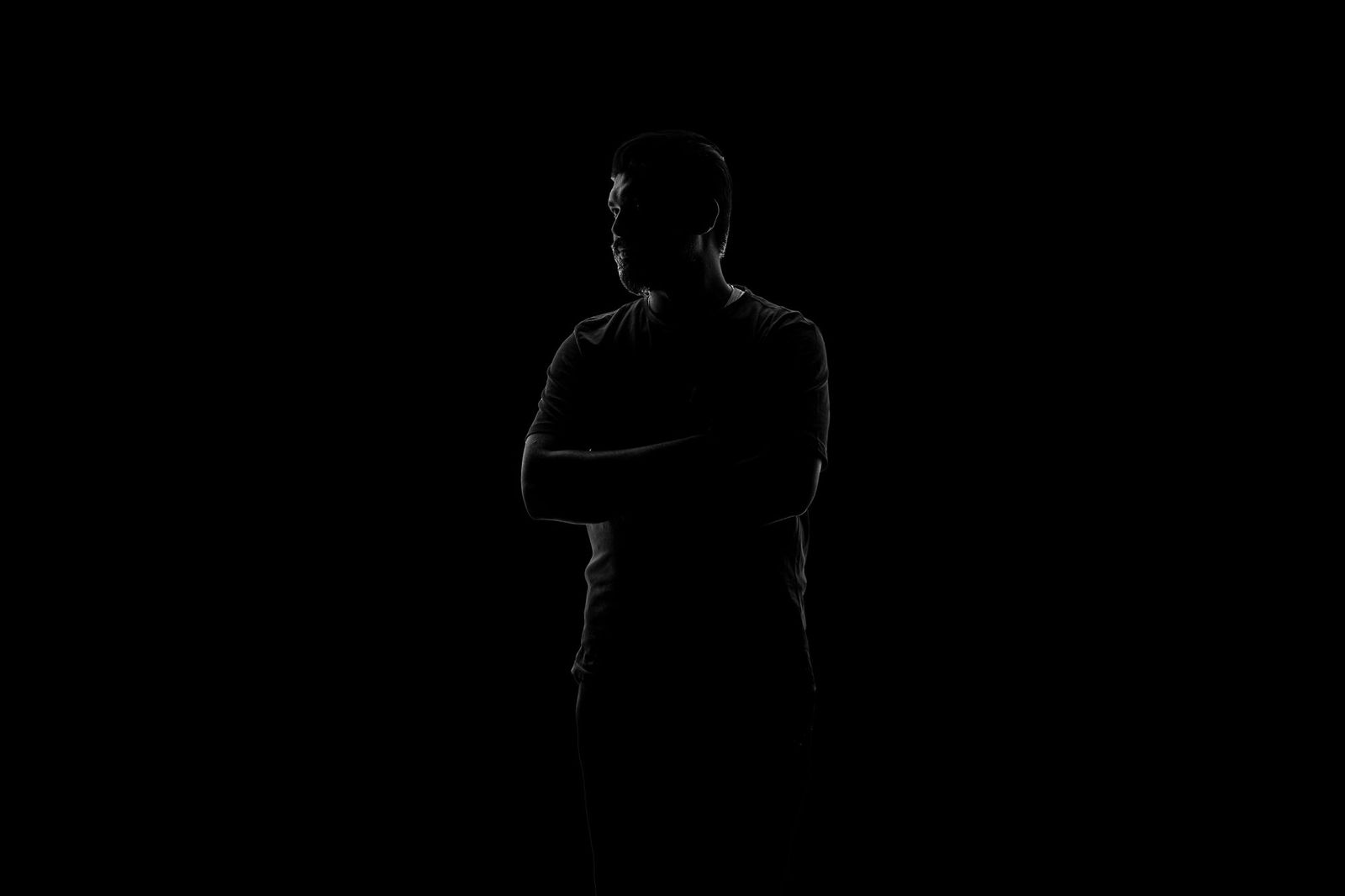 A silhouetted figure standing with arms crossed against a plain dark background, face turned to the side