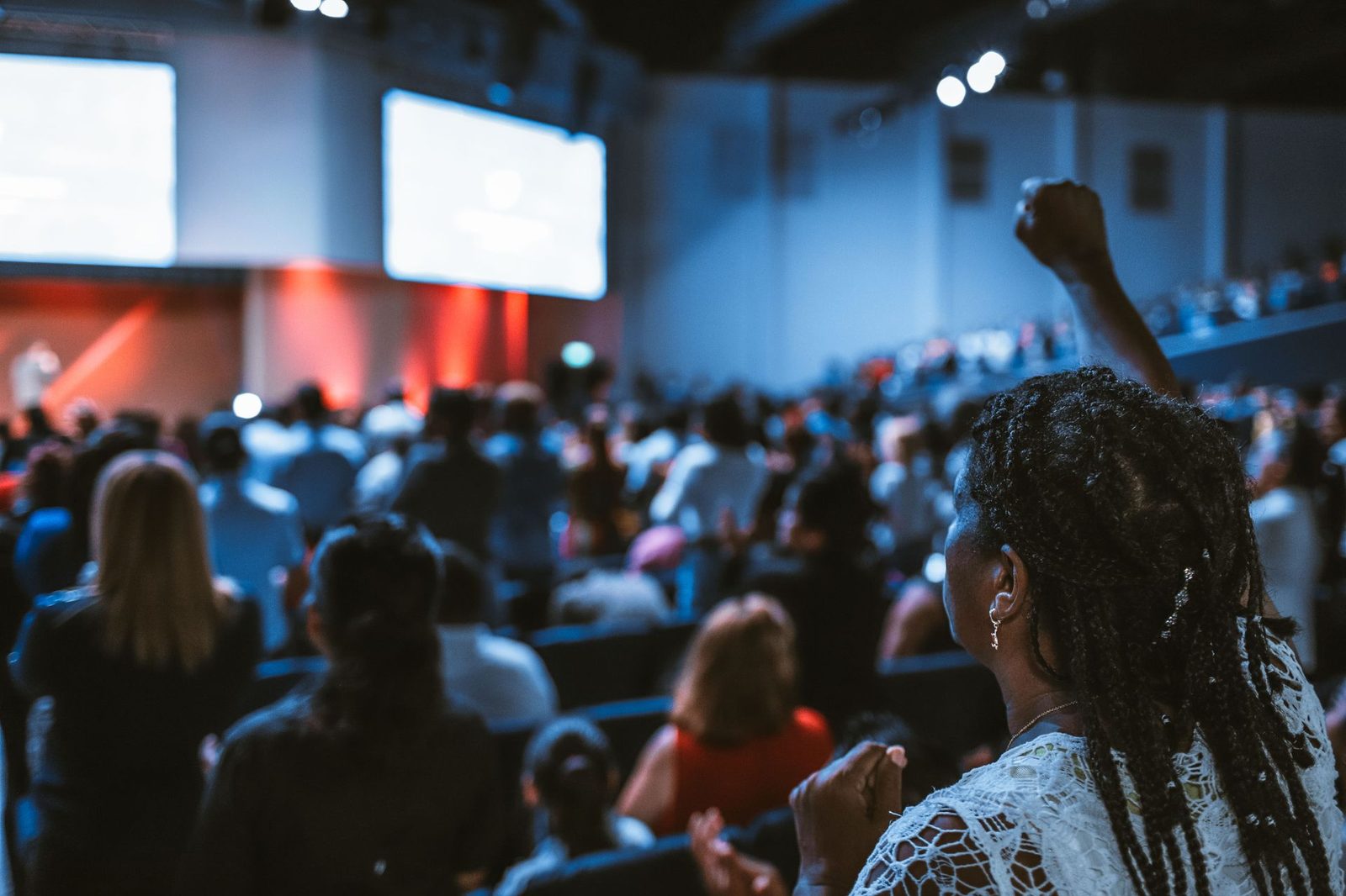 A packed auditorium seen from the back, audience facing a lit stage with presentation screens
