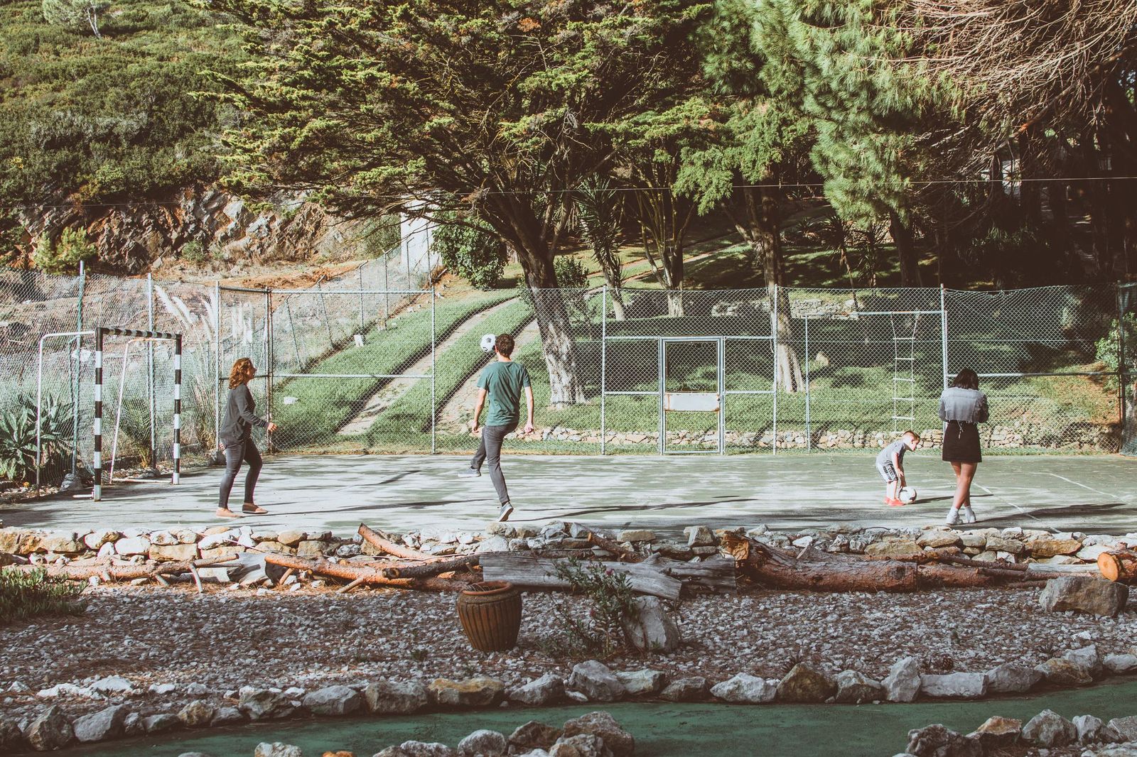 Four people kicking a football around a fenced outdoor court surrounded by trees