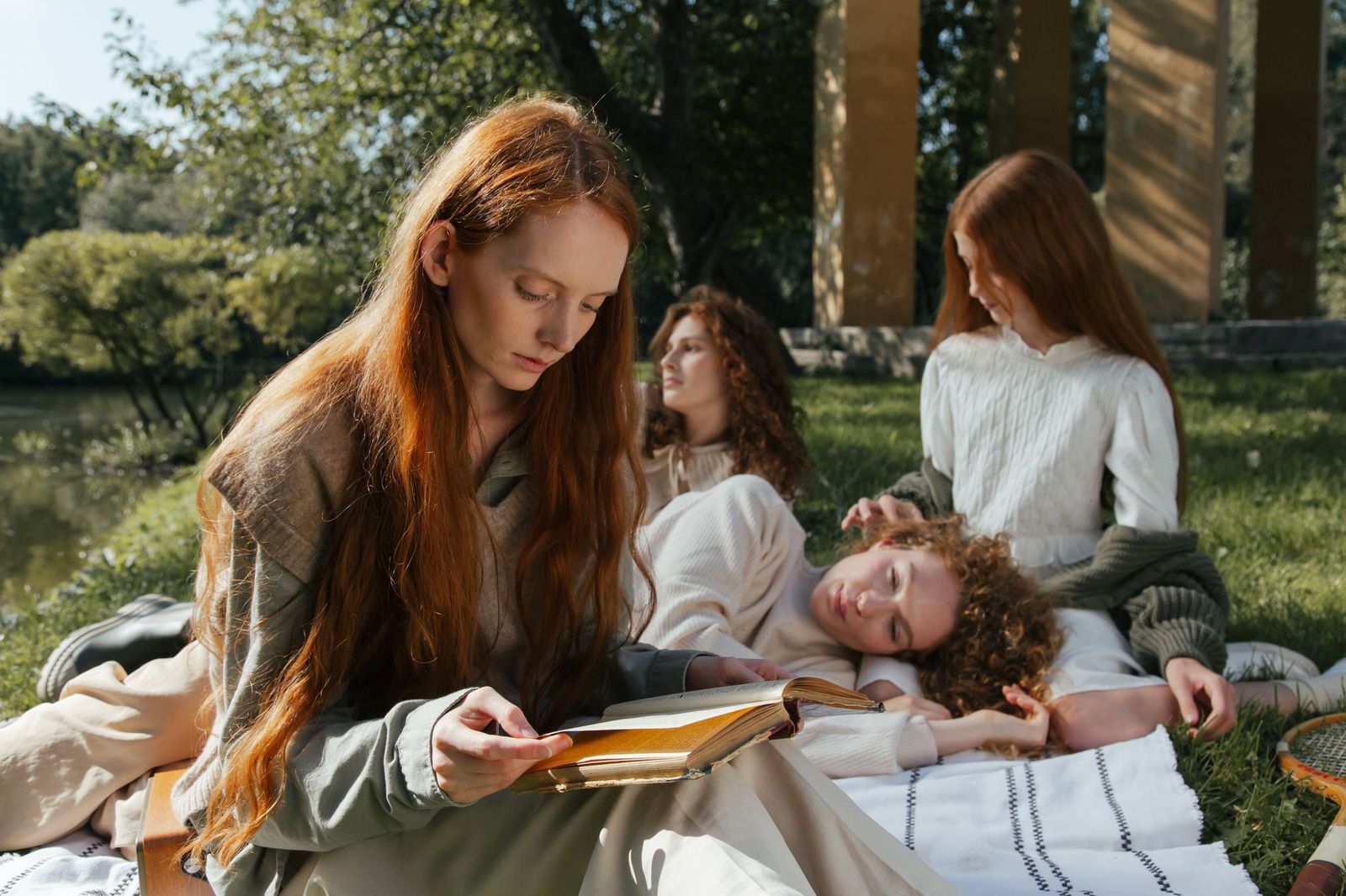 Four friends sitting on blankets on the grass in a park, one reading an old book aloud while another rests beside her