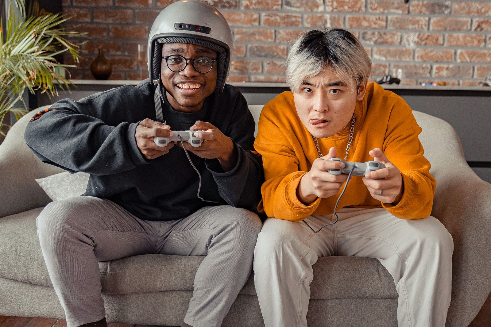Two friends sitting on a sofa playing a console game with wired controllers, one of them wearing a motorcycle helmet, in front of a brick wall