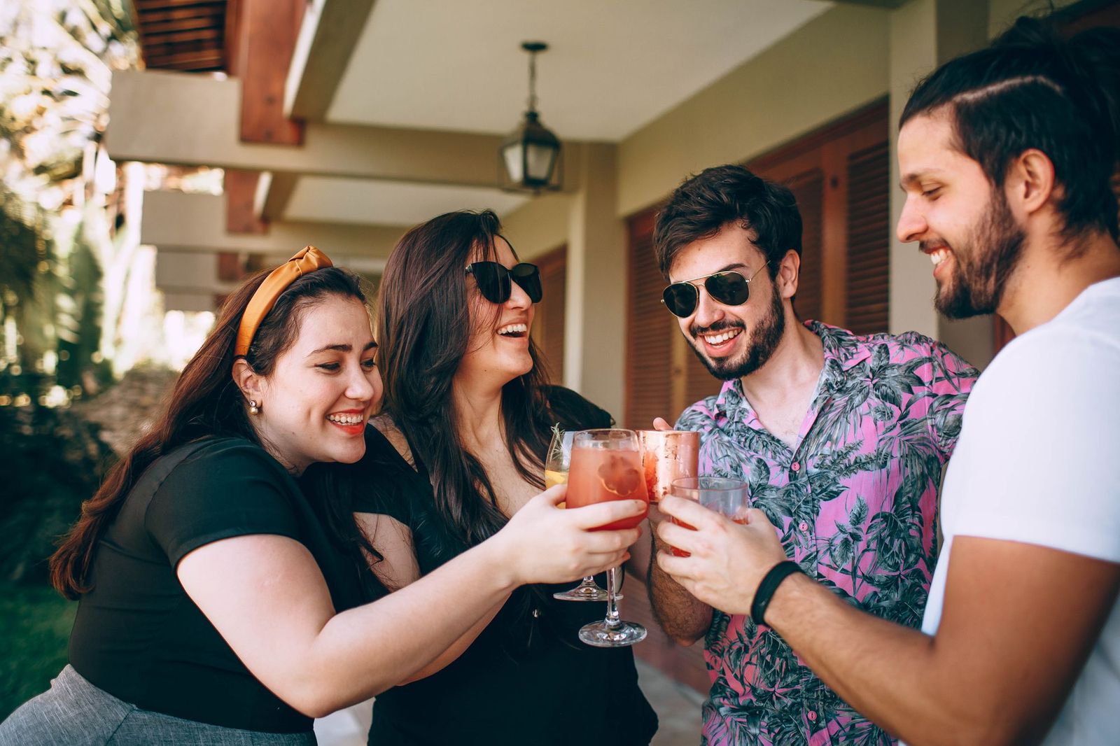 Four friends on a sunlit porch leaning in to clink glasses of pink and orange drinks together, all laughing