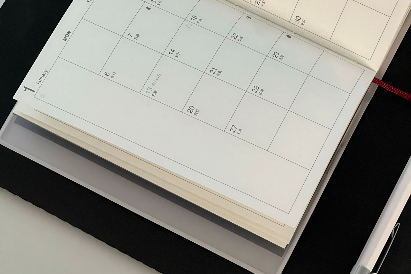 An open paper monthly planner on a dark desk with every date square left blank, a red ribbon marker and a pen beside it