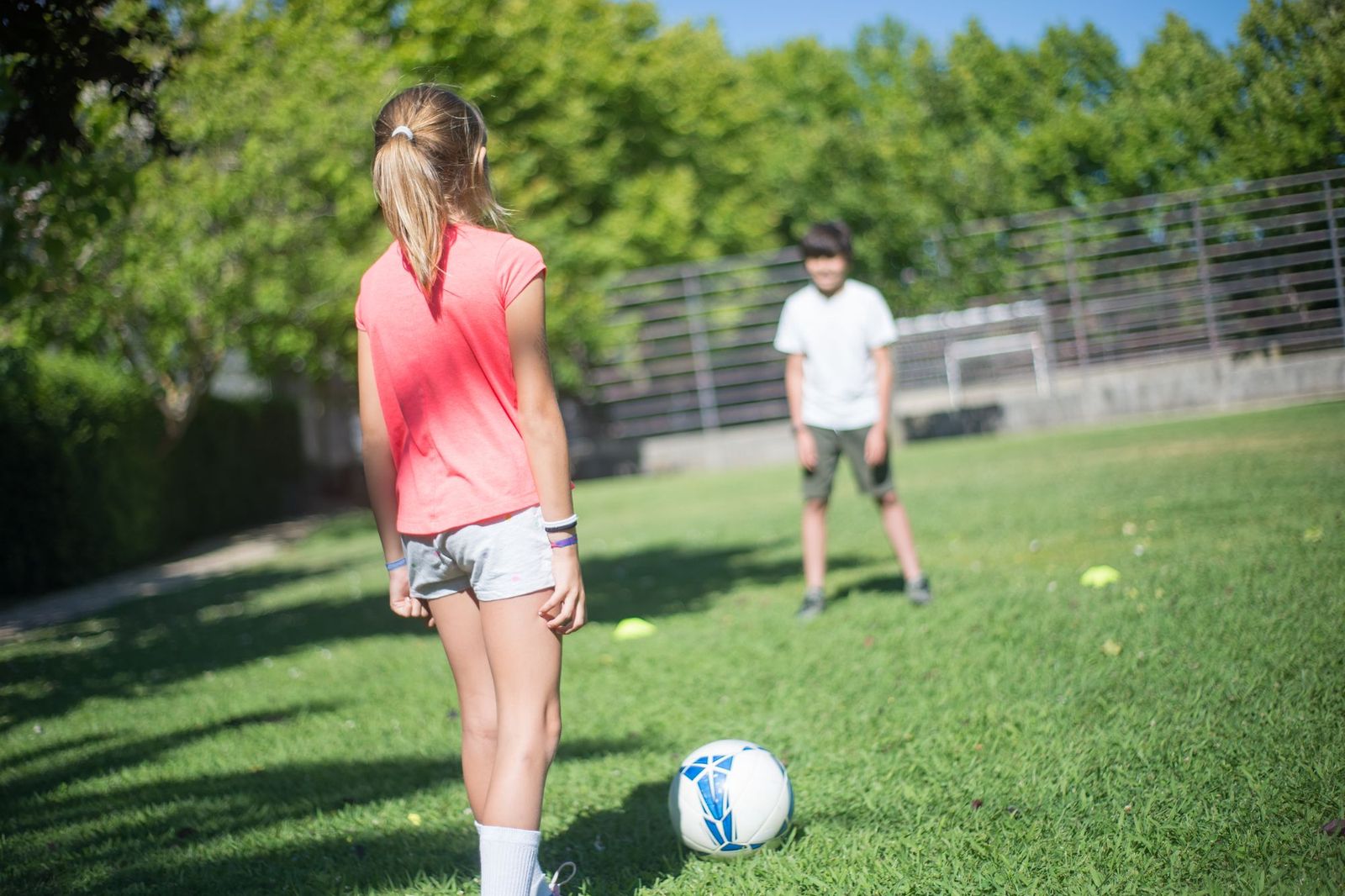 Two young people playing football on a sunny grass pitch, one standing over the ball and the other waiting in front of a goal