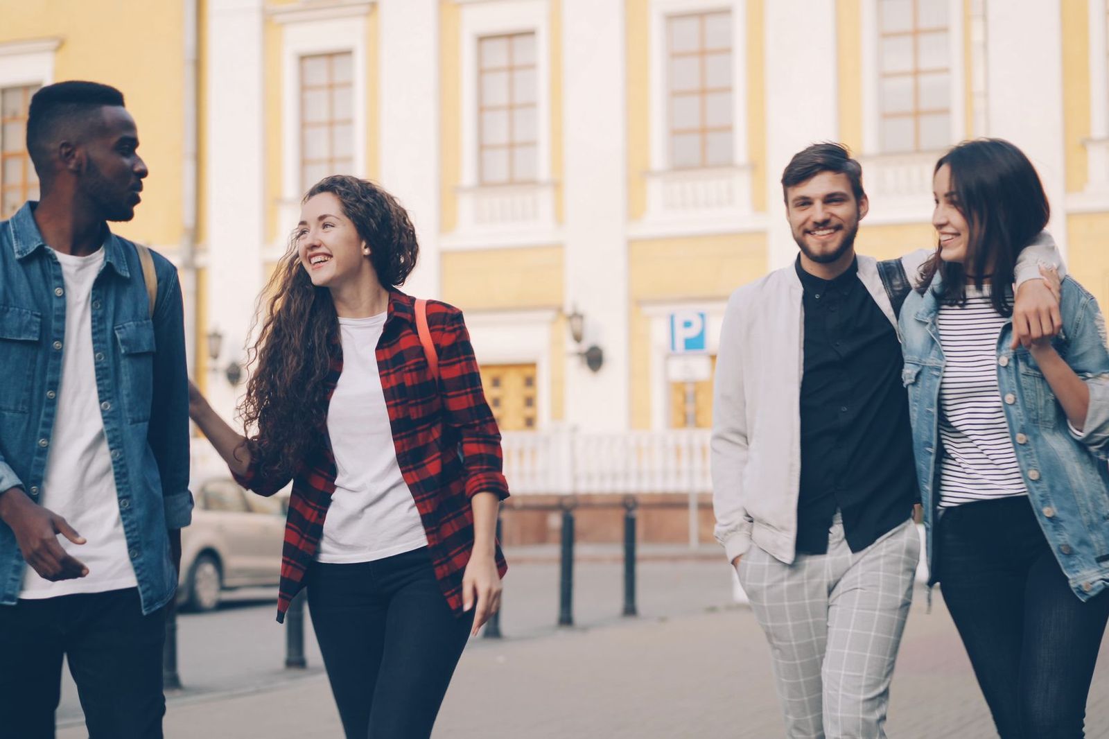 Four friends walking together along a city street in front of a yellow building, talking and laughing