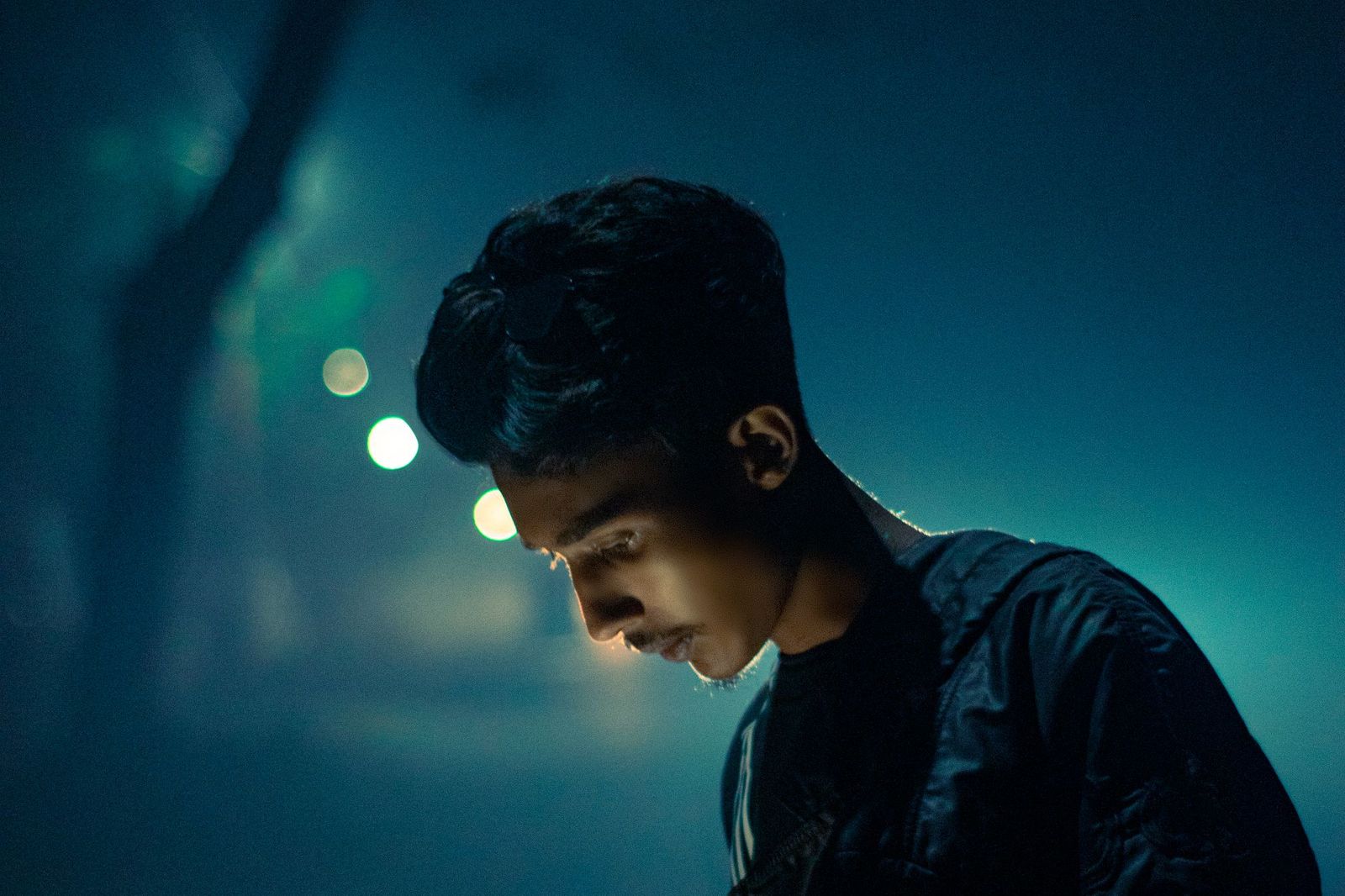 A young man walking alone at night, head down, lit by cool blue light with soft bokeh from streetlights behind him