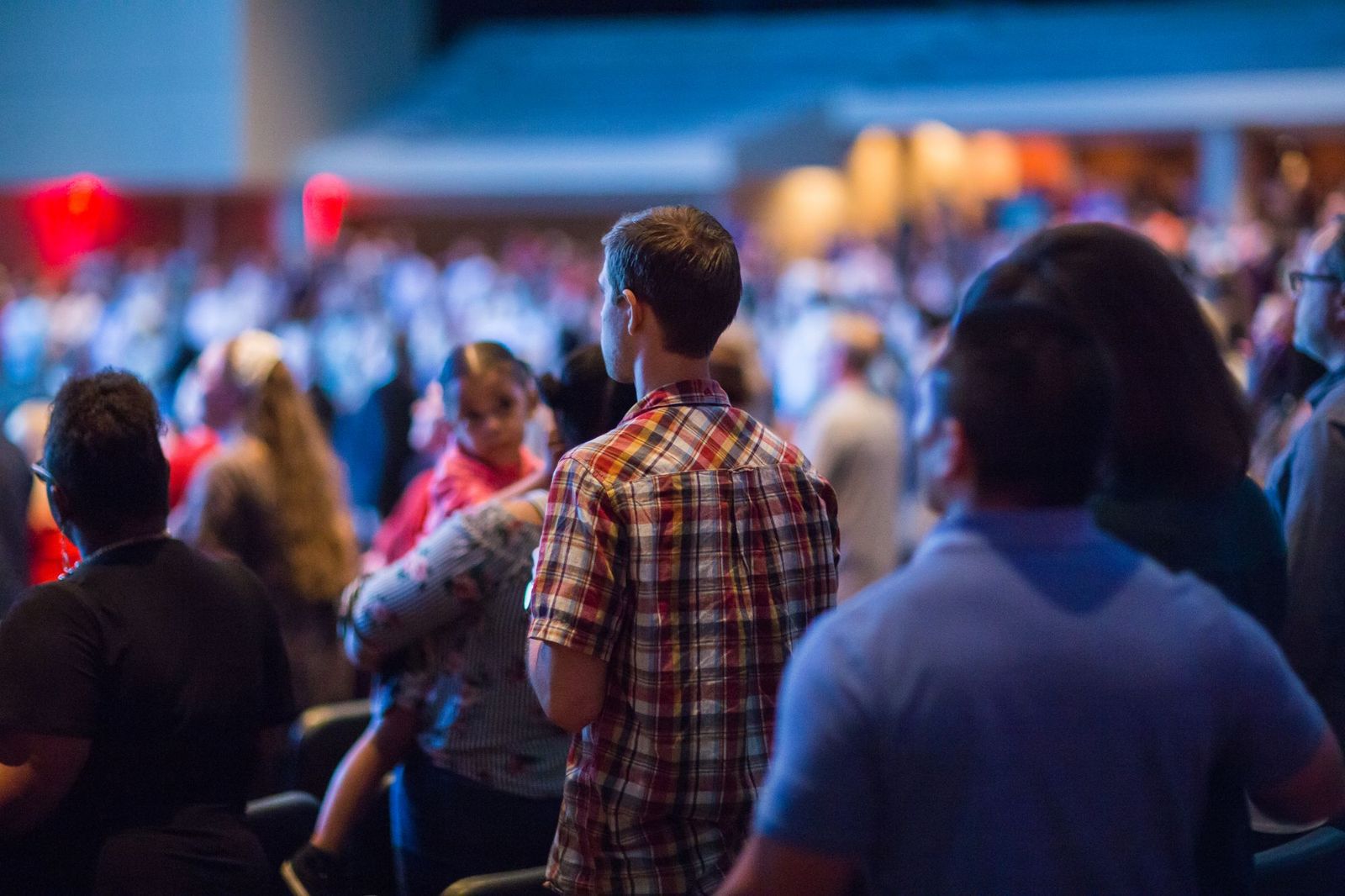 A crowd of concertgoers standing shoulder to shoulder in a dim venue lit with red stage lights, a man in a plaid shirt seen from behind