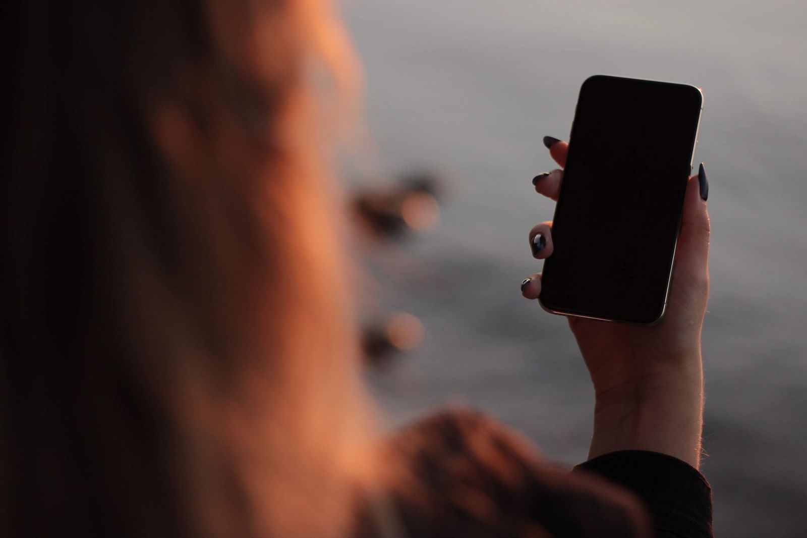 A person holding a smartphone with a dark screen up near their face at dusk, standing by water with two ducks visible in the blurred background