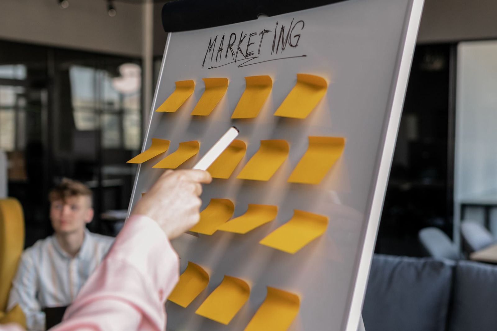 A person in a pink shirt holding a marker and pointing at a flipchart headed Marketing covered in rows of yellow sticky notes, with a colleague watching from a chair behind