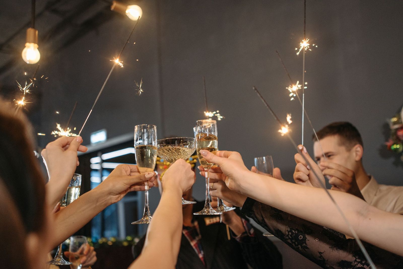 A group of people raising champagne glasses together in a toast while holding lit sparklers, under warm bulb lighting at an indoor party