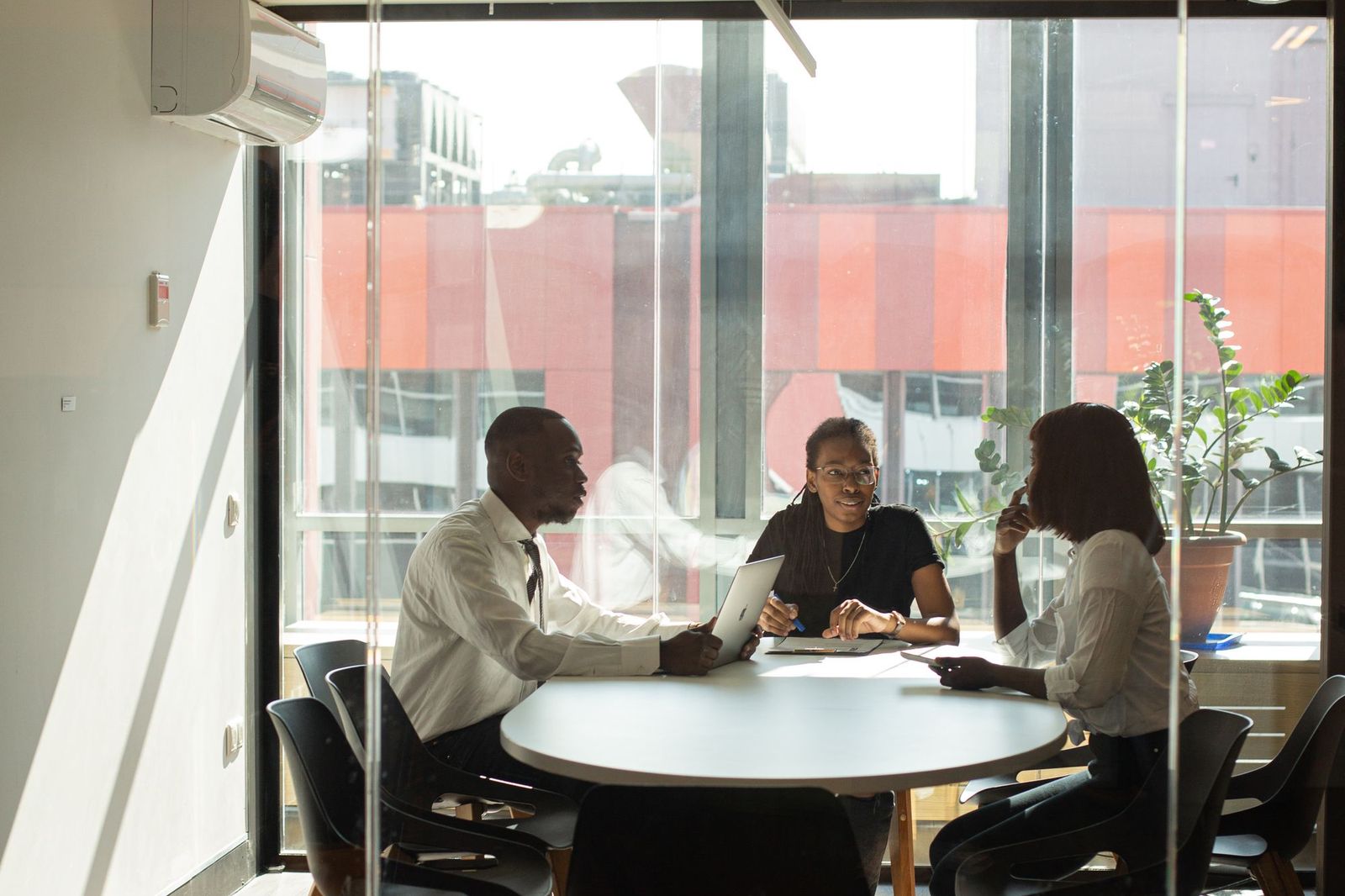 Three colleagues sitting around a small round table in a glass-walled meeting room, talking, with a laptop and notebook between them and city buildings visible through the window