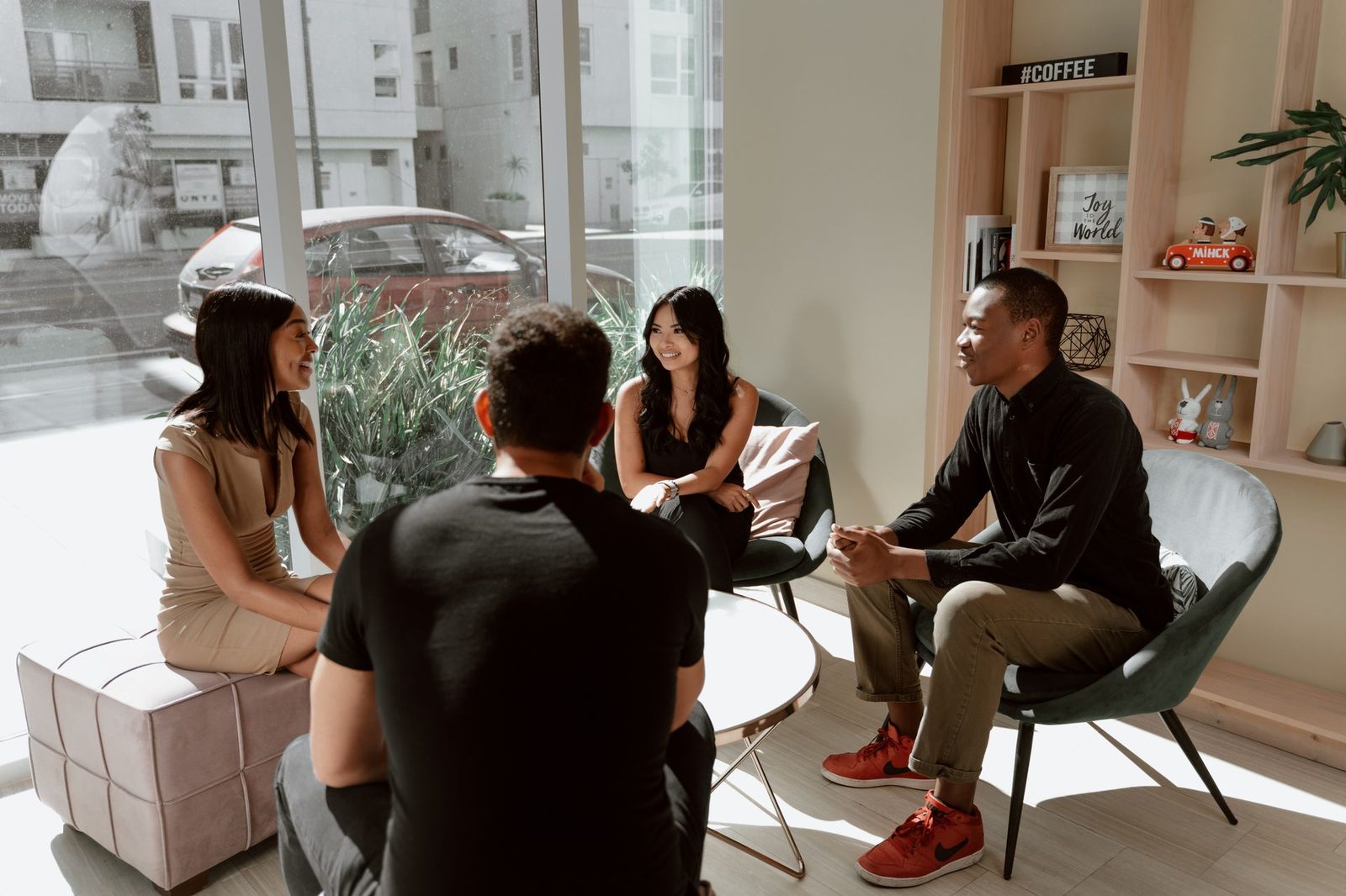 A small group of people sitting around a low table in a bright café, mid-conversation, laptops and cups between them