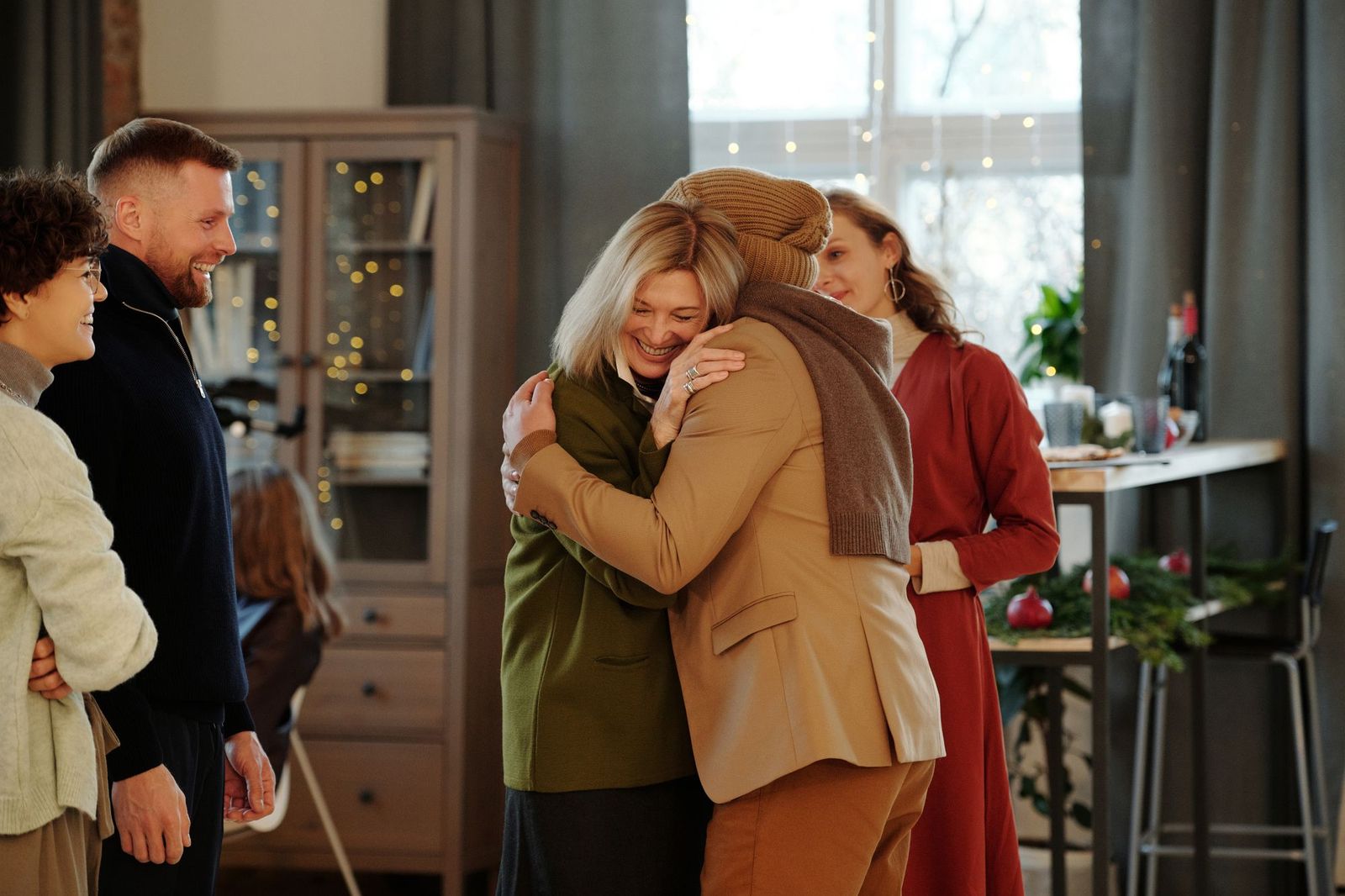 A warm group of friends embracing at a gathering, several people smiling and greeting each other in a living room decorated for the holidays