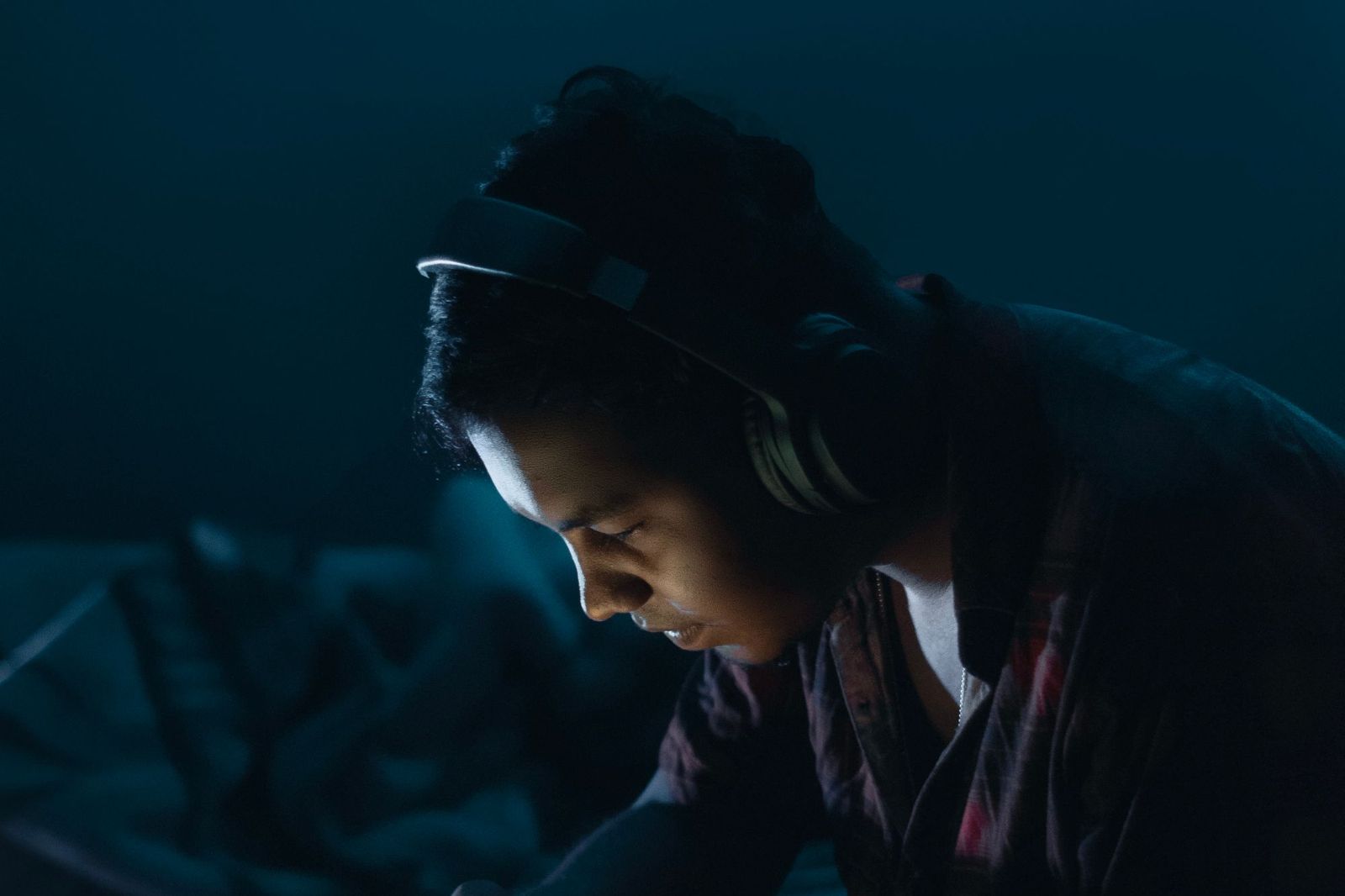 A person alone at night, lit by the cold glow of a phone or laptop screen, headphones on, leaning forward in concentration