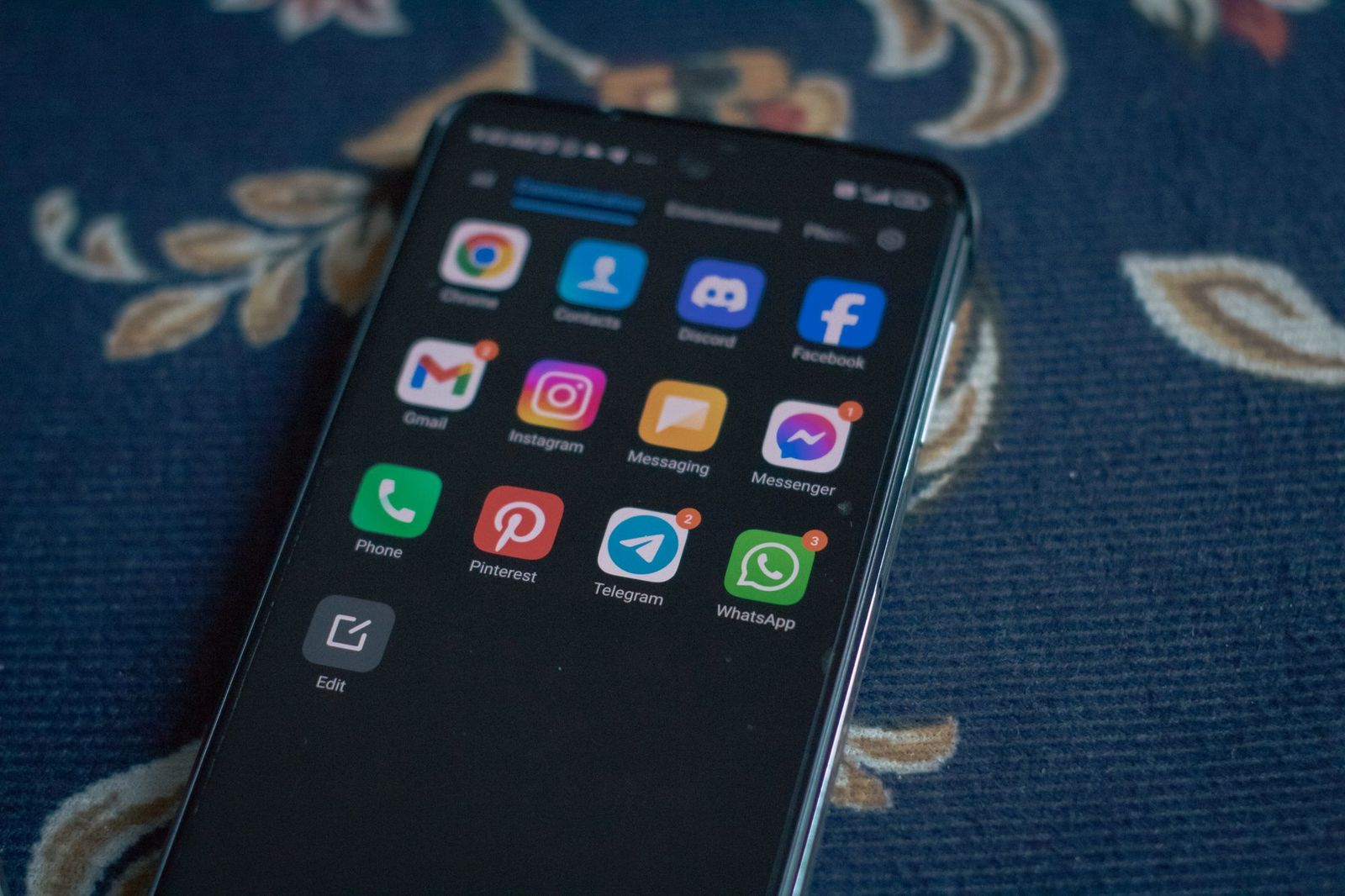 A smartphone lying face up on a patterned rug, showing a home screen with Discord, Facebook, Messenger, Telegram and WhatsApp app icons