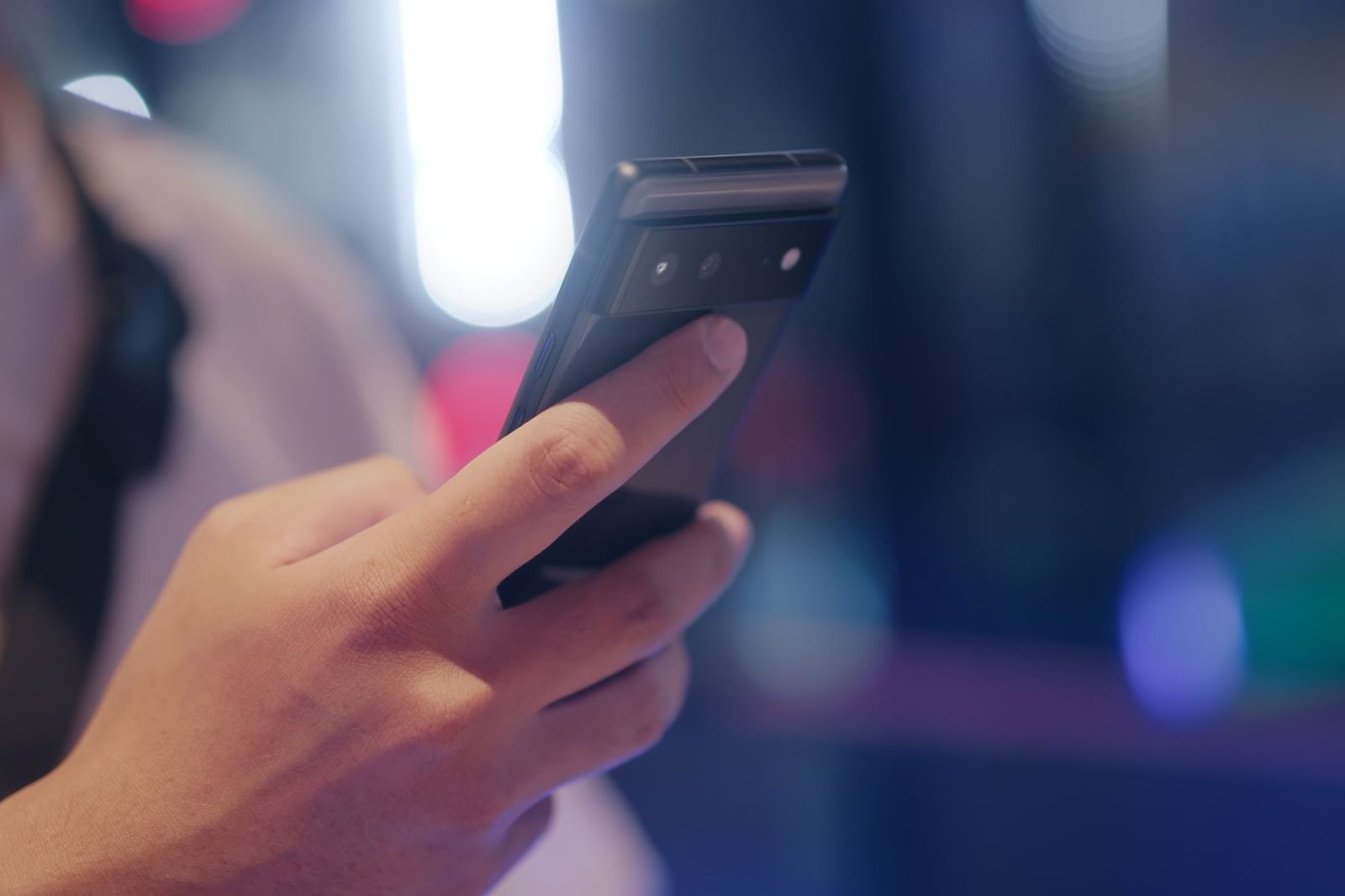 A hand holding a smartphone with a dark screen up close, illuminated by soft colourful bokeh lights at night