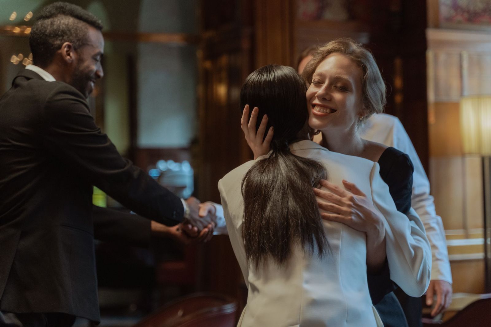 A man and woman shaking hands and embracing warmly in a wood-panelled room lit by warm lamps, other guests nearby