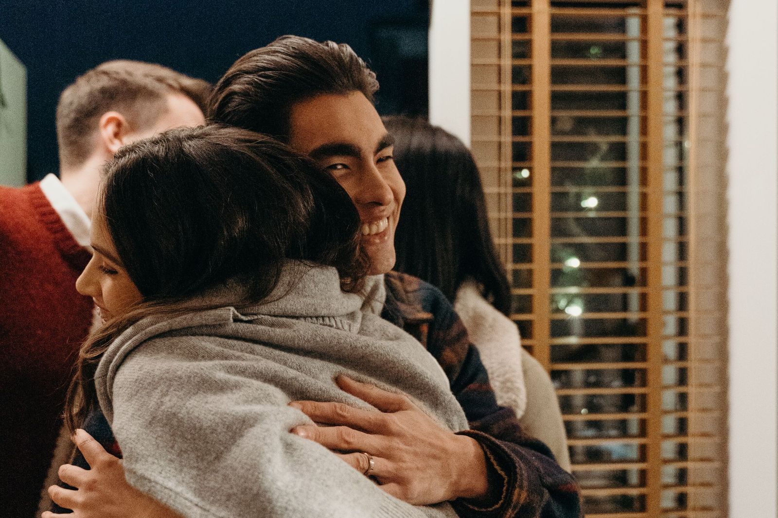 A man and woman embracing warmly in a doorway at an evening gathering, with other guests visible in the background