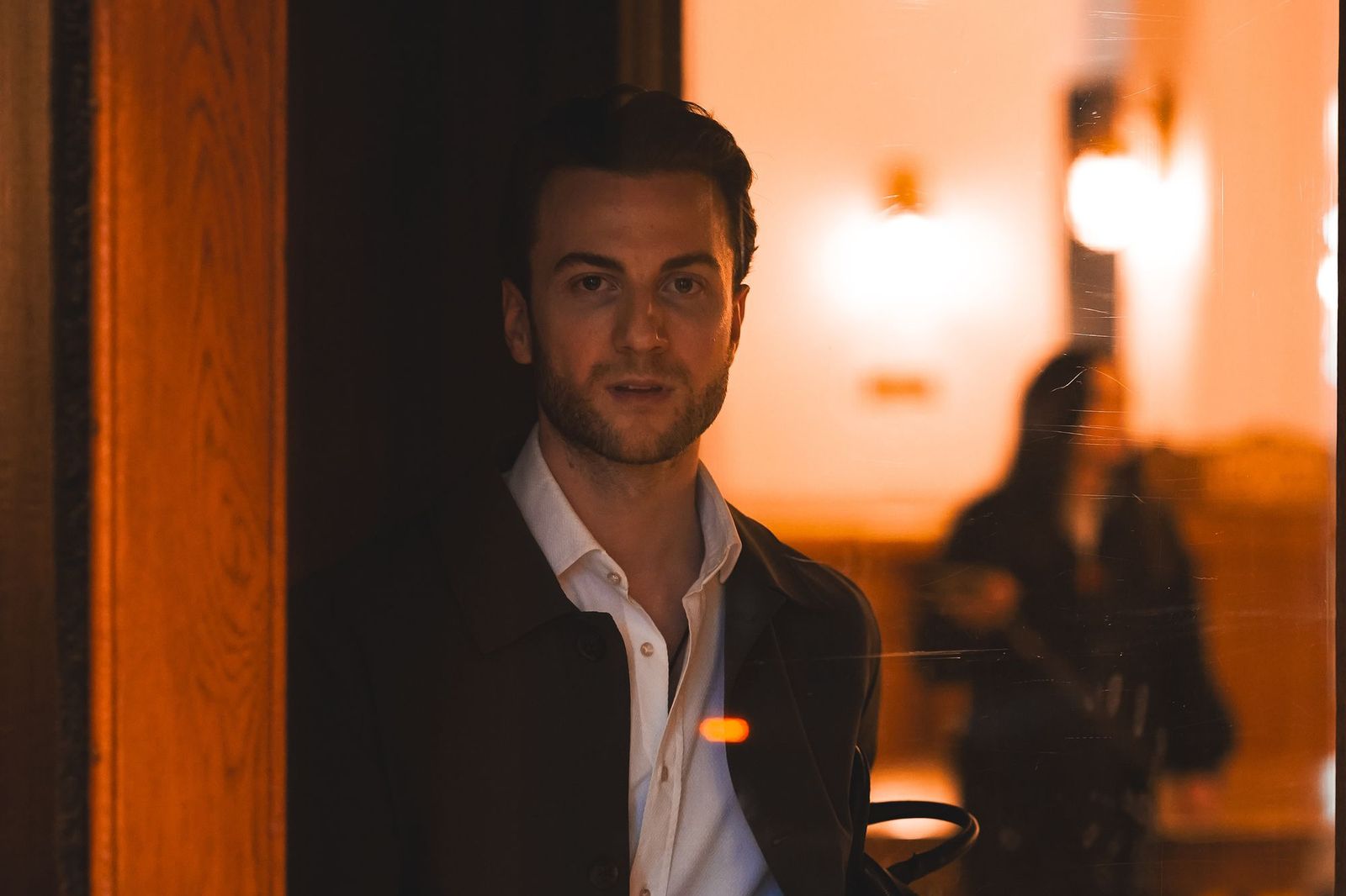 A man standing in a doorway at night looking toward the camera, warm orange light and a blurred figure visible behind him