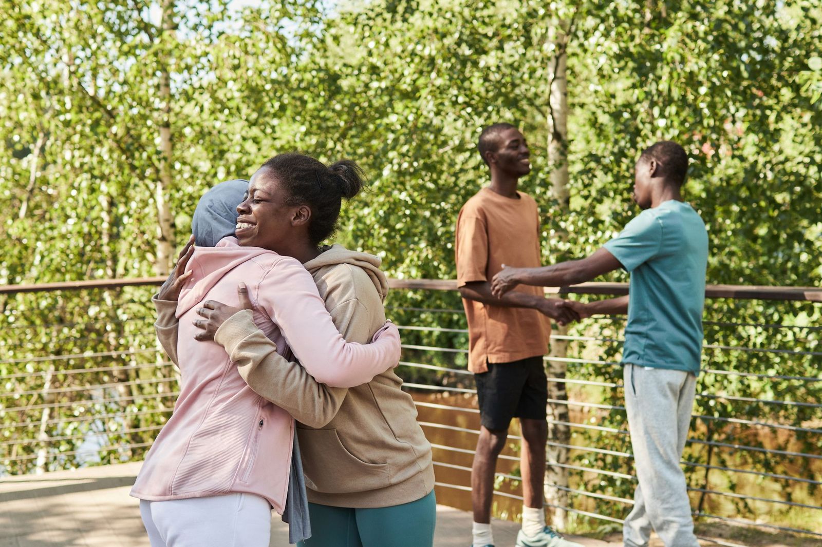 Two friends embracing warmly outdoors while two other people shake hands beside them on a bridge lined with trees