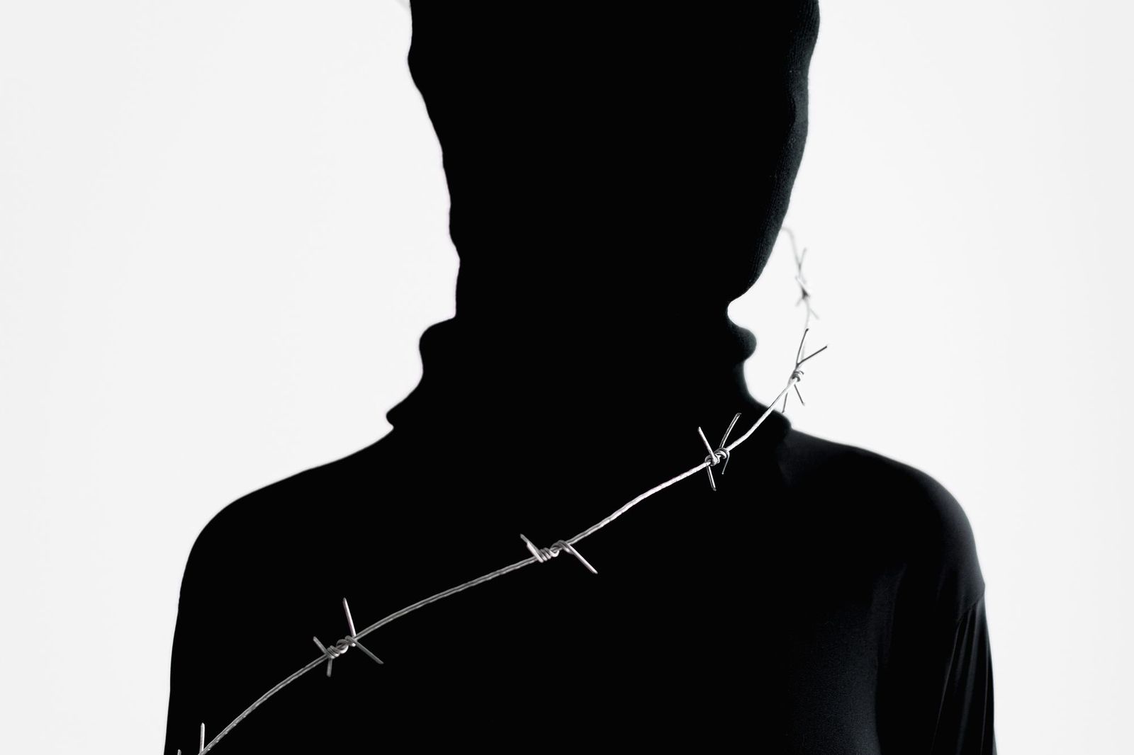 A dark, minimalist photograph of a person's silhouette against a plain white background, a strand of barbed wire crossing diagonally in front of their neck and shoulder