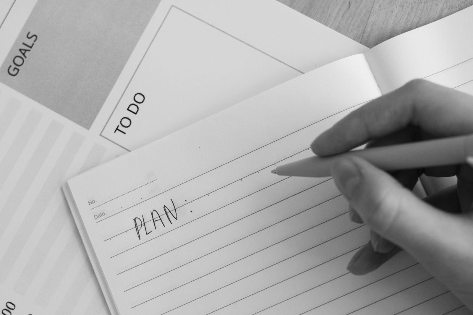 A hand holding a pen writing the word PLAN on the first line of a lined notebook, with blank goals and to-do sheets fanned out beside it