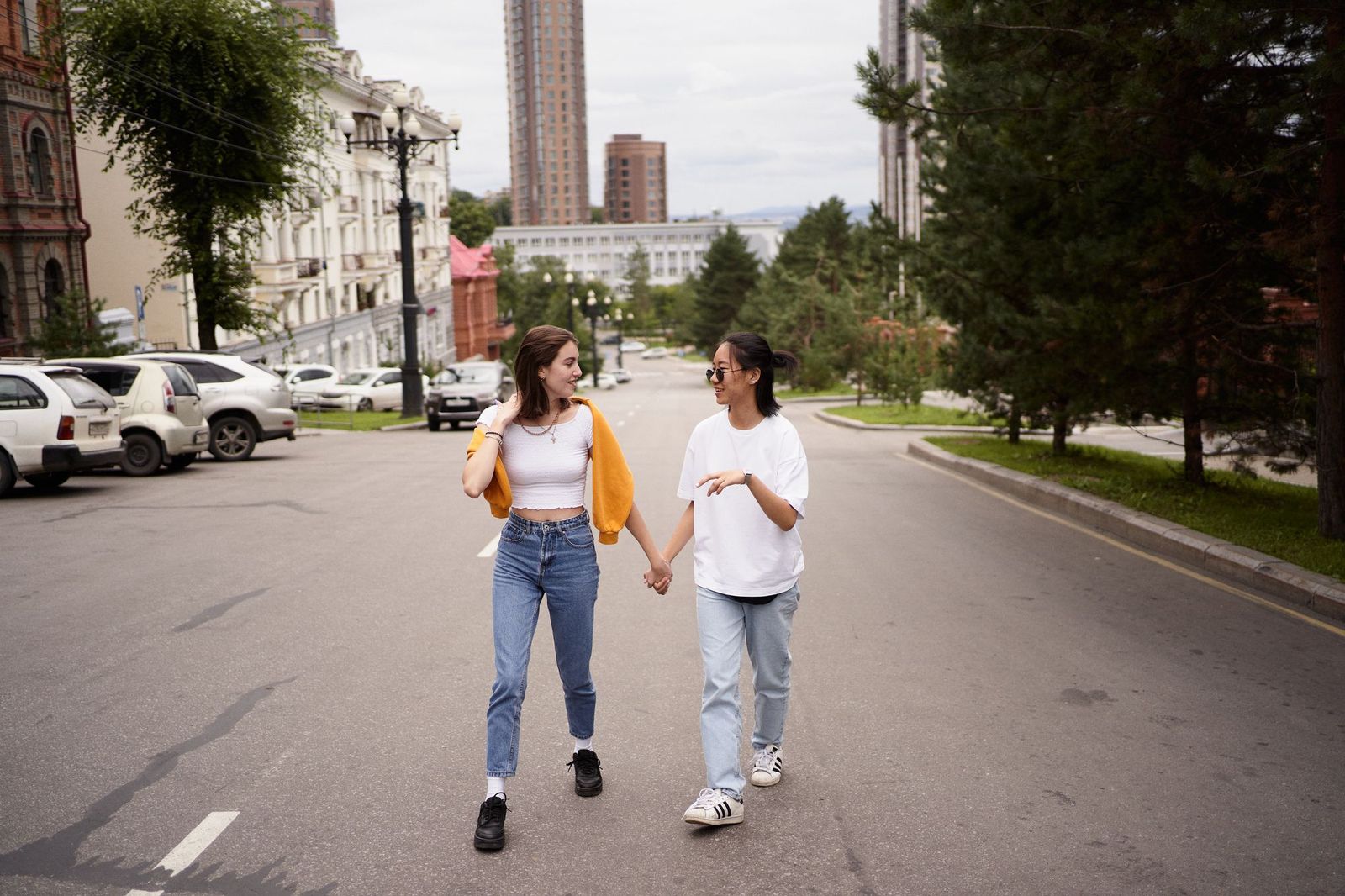 Two young women walking down the middle of a quiet city street holding hands and talking to each other