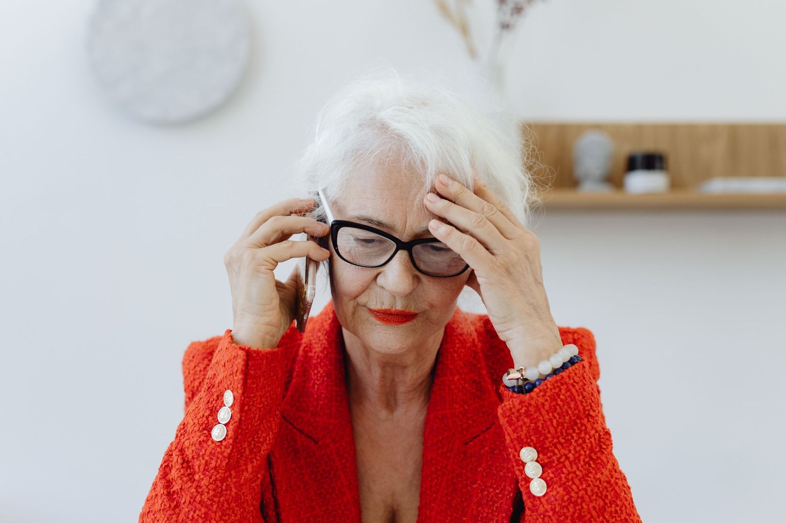 An older woman in a bright red jacket holding a phone to her ear with one hand pressed to her forehead, looking strained