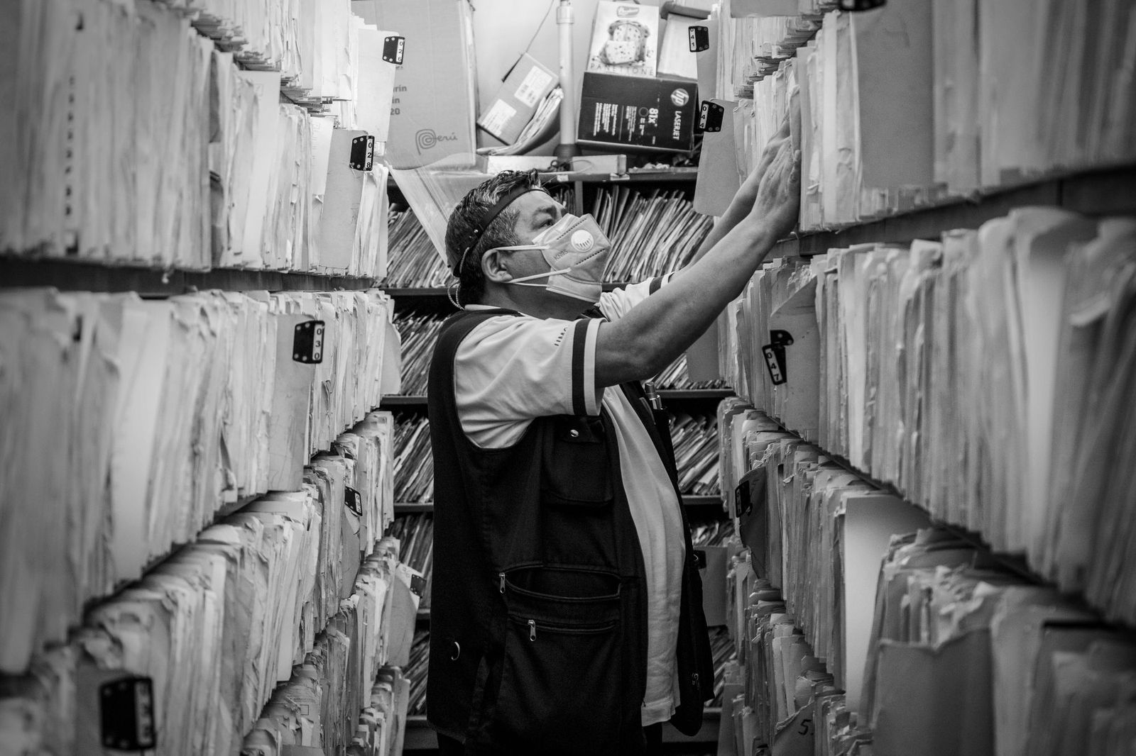 A worker in a mask reaching up to pull a folder from a towering aisle of densely packed paper files stretching in both directions