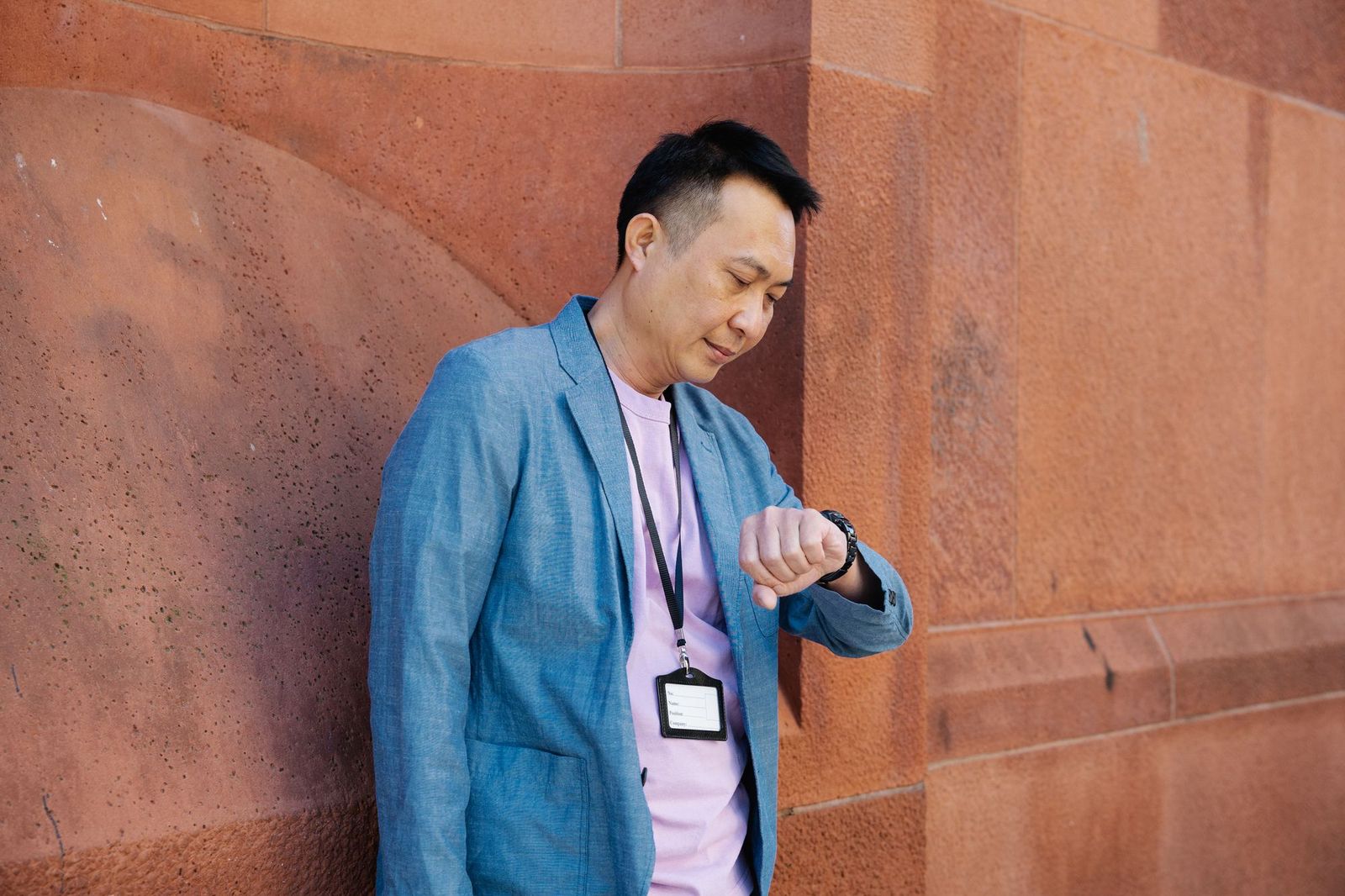 A man in a blazer with a lanyard leaning against a terracotta wall, checking the time on his watch