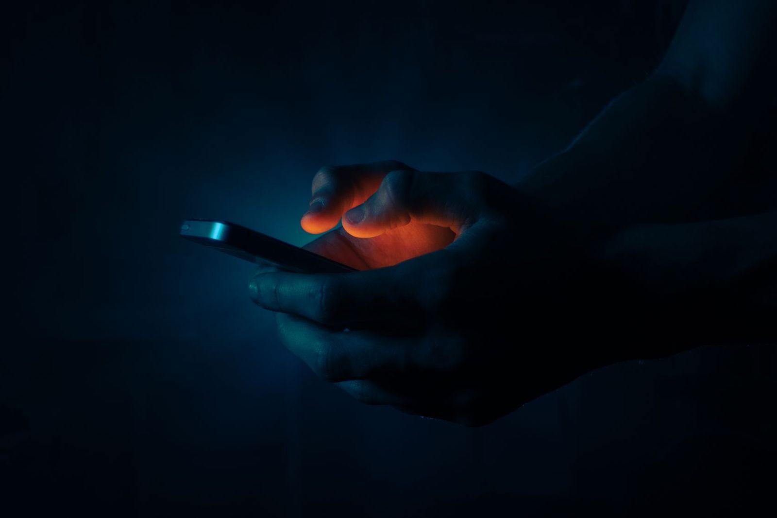 A close-up of a hand holding a smartphone in a dark room, the screen glowing blue and orange against the person's fingers