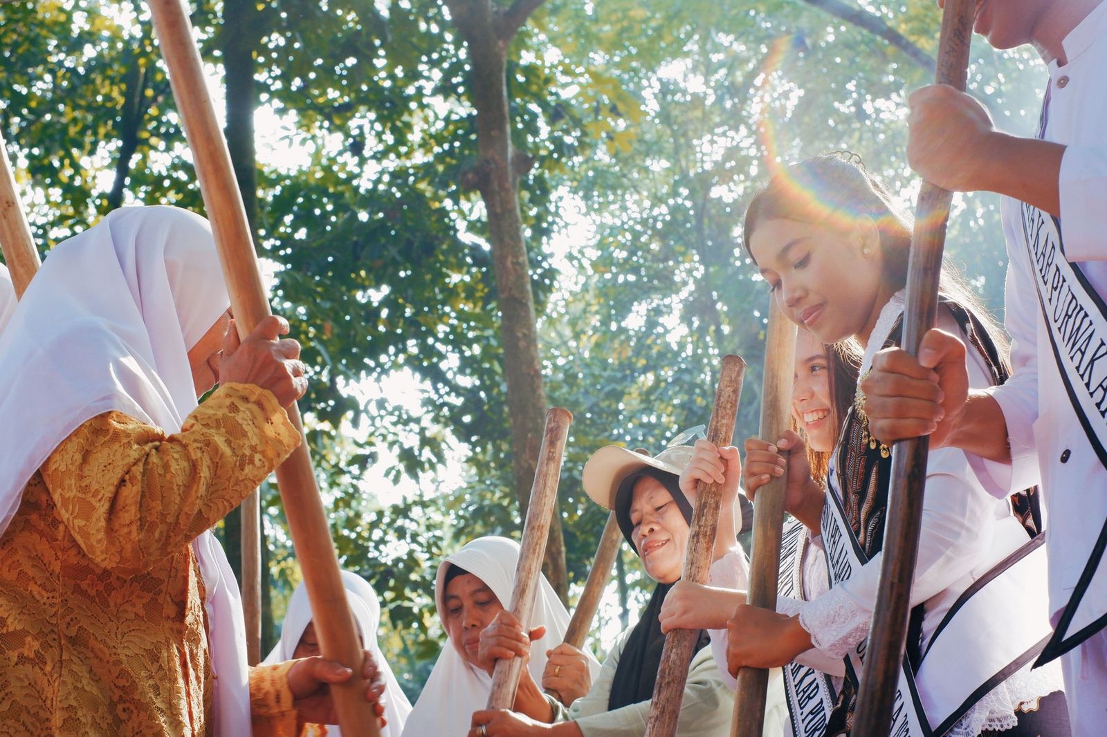 A group of people outdoors under trees holding long wooden poles together, smiling as they take part in a community gathering