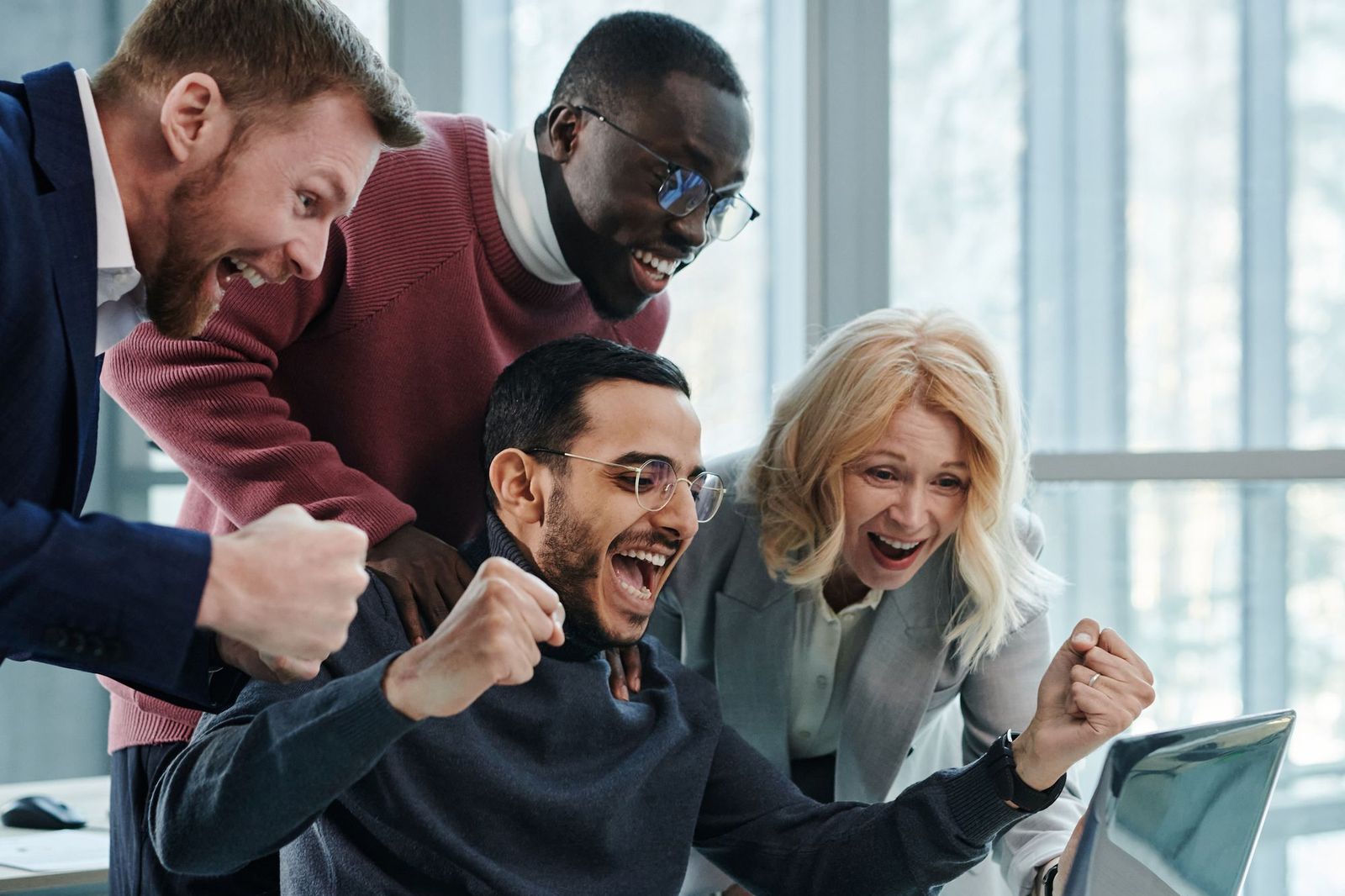 Four colleagues crowded around a laptop in a bright office, punching the air and cheering at something on the screen