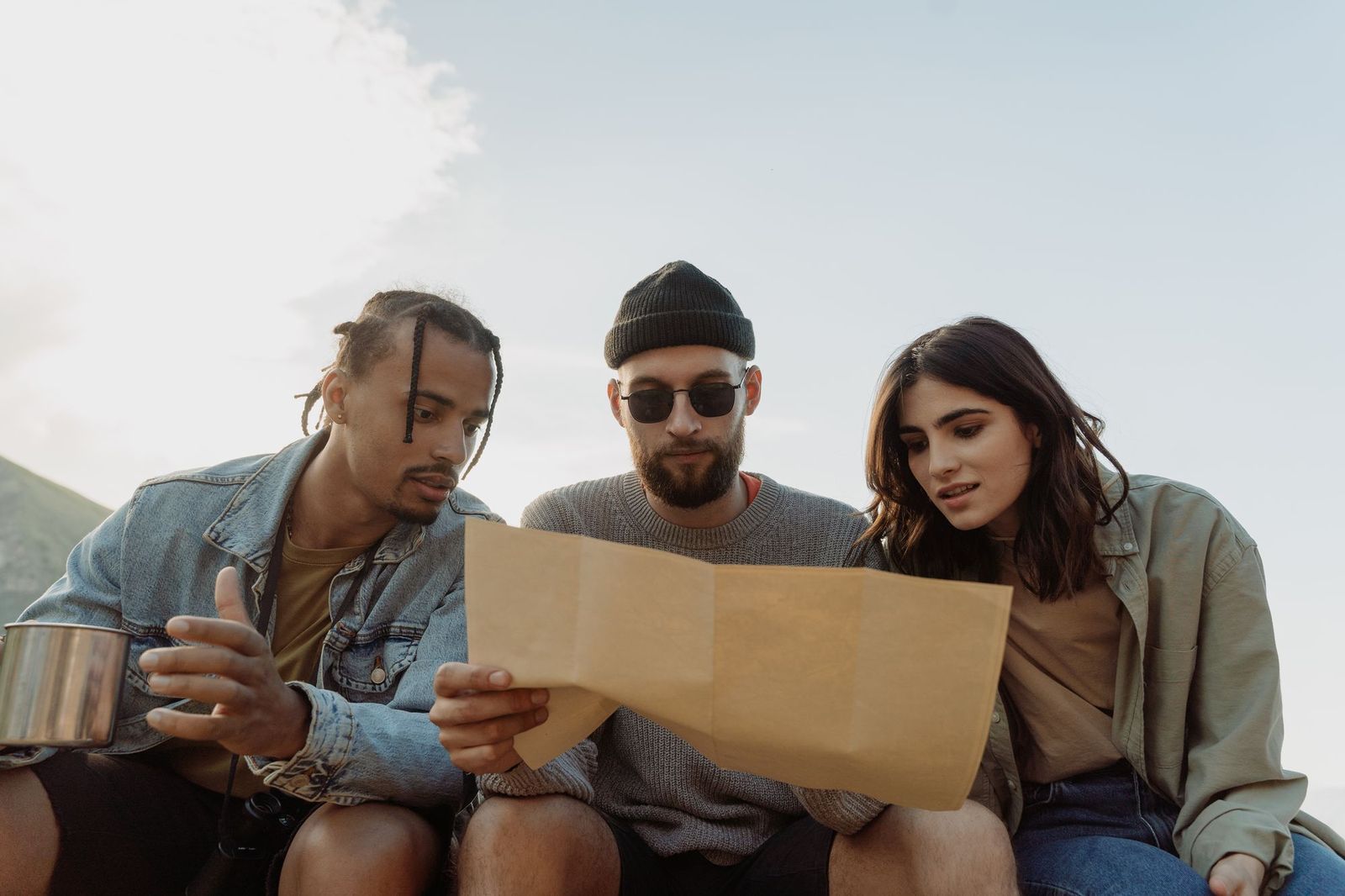 Three friends sitting outdoors against a bright sky, leaning in together over an unfolded paper map