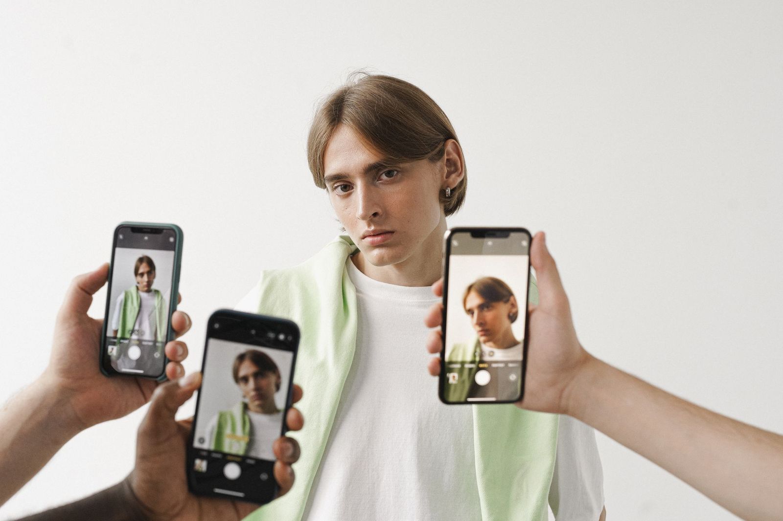 A young man in a white t-shirt stands against a plain wall while three hands hold up phones in front of him, each screen showing a slightly different camera view of the same person