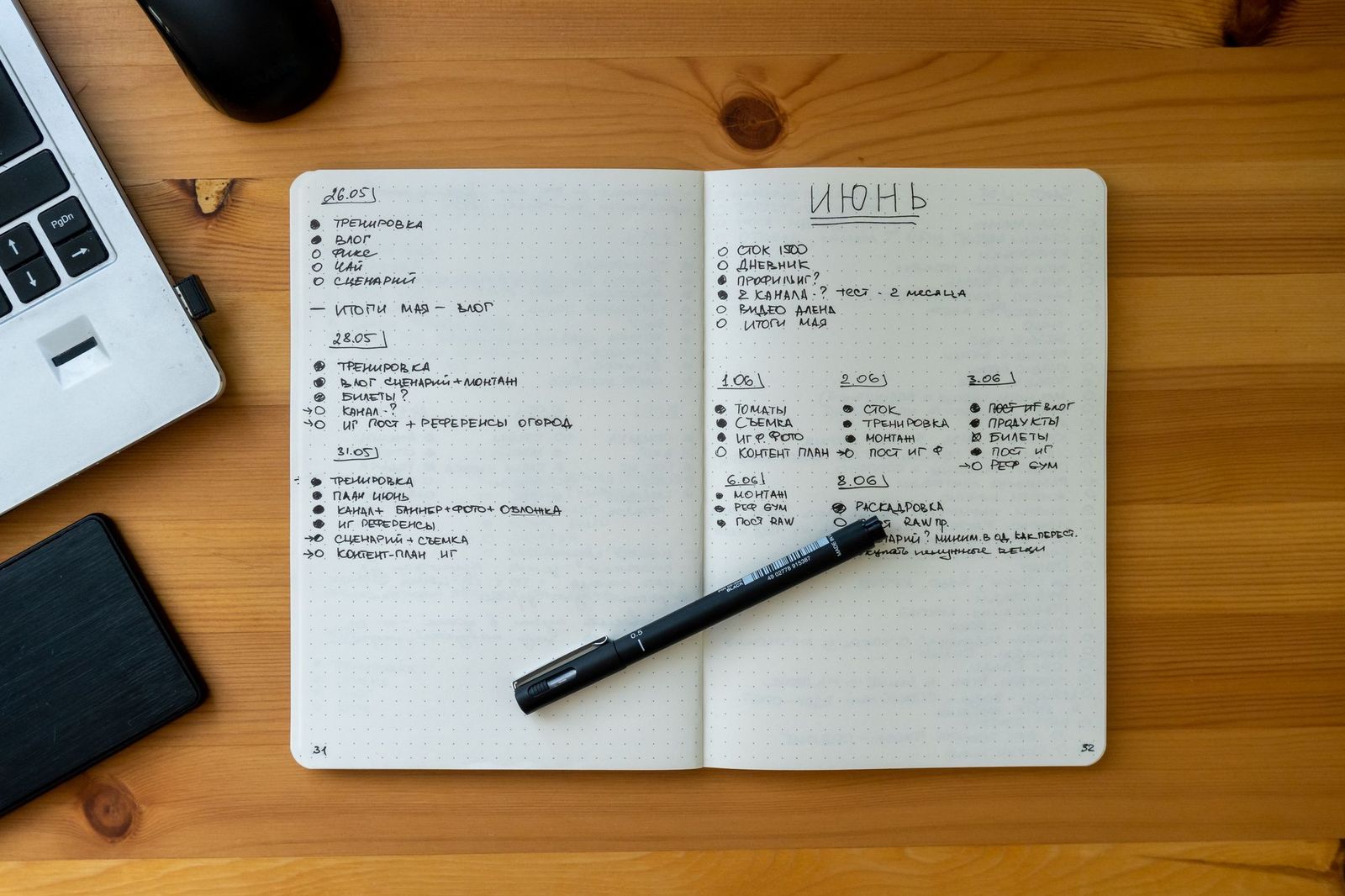An open dot-grid notebook on a wooden desk covered in handwritten dated to-do lists, a fineliner pen resting across the pages beside a laptop