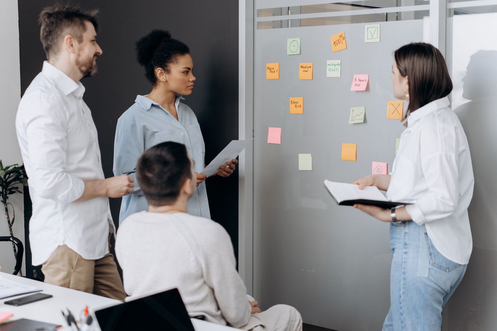 Four colleagues standing and sitting around a glass wall covered in handwritten sticky notes in an office, two of them holding papers and a notebook