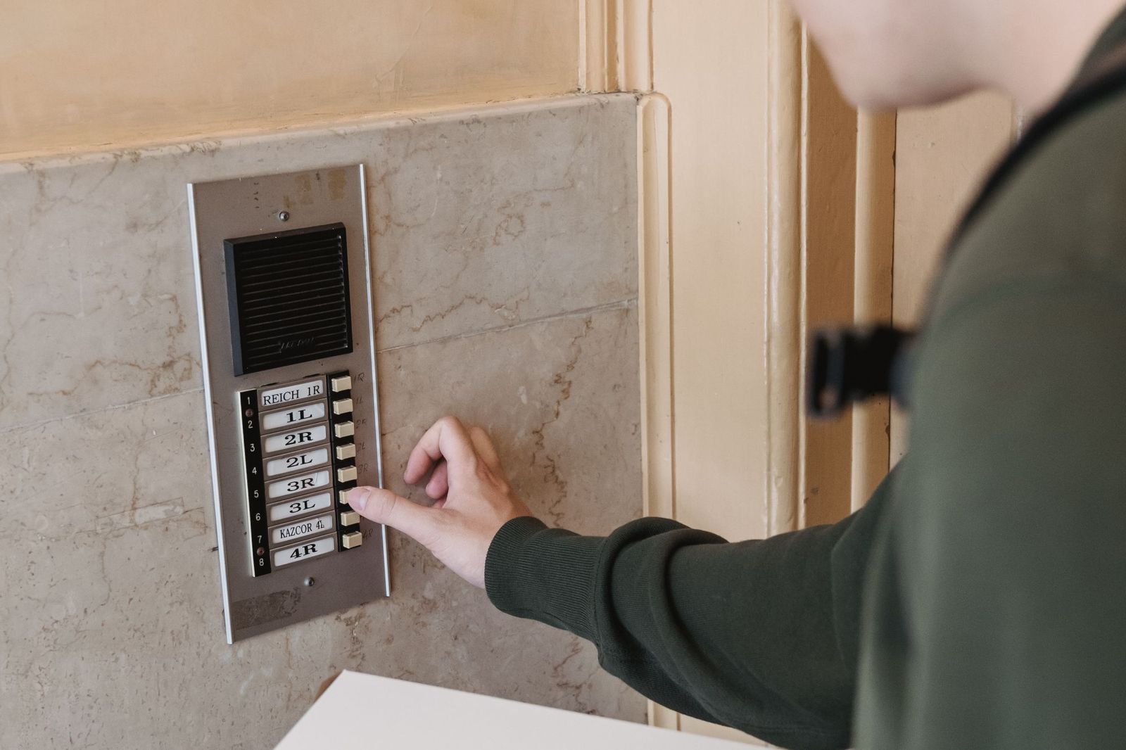 A hand pressing one of eight numbered buzzer buttons on a metal door intercom panel set into a marble entrance wall
