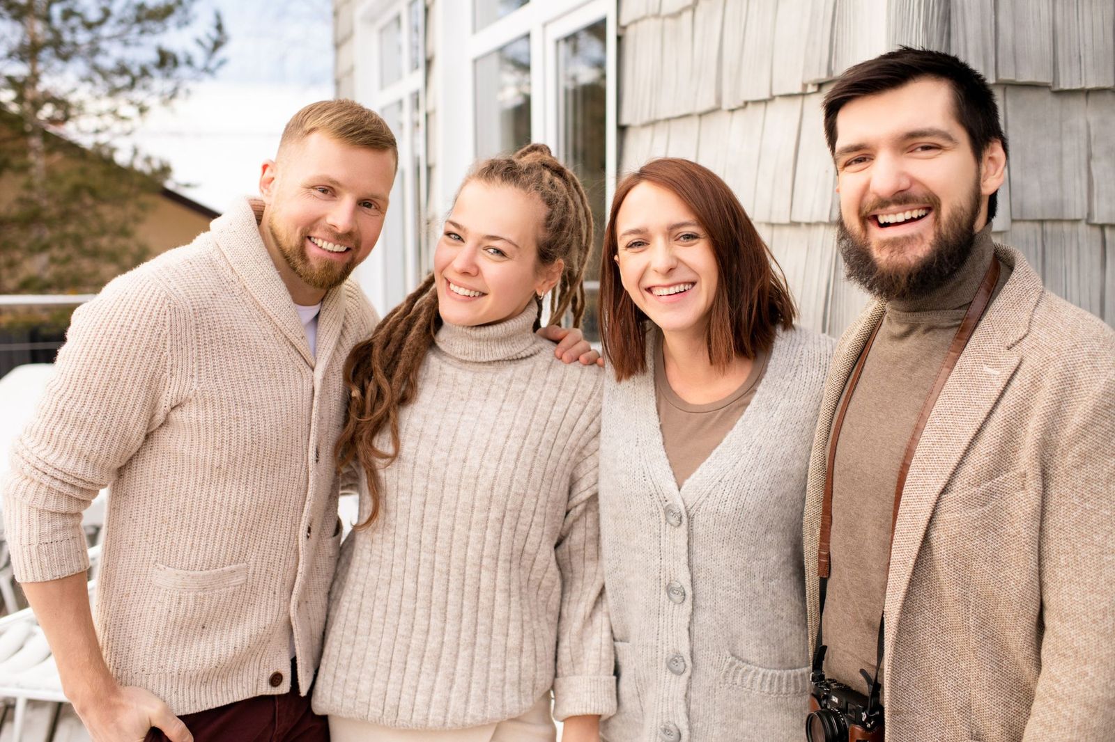 Four friends in knitted jumpers and coats standing shoulder to shoulder outside a house, smiling at the camera
