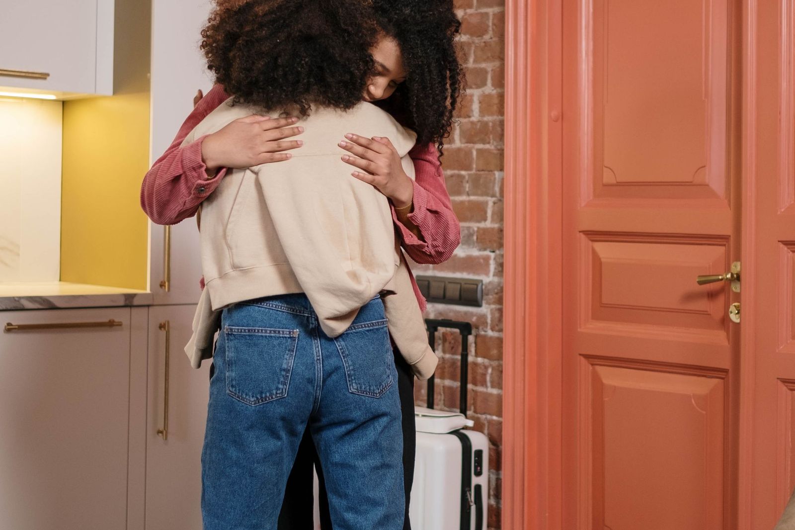 Two people hugging in a doorway of a flat, a wheeled suitcase standing beside them next to a brick wall
