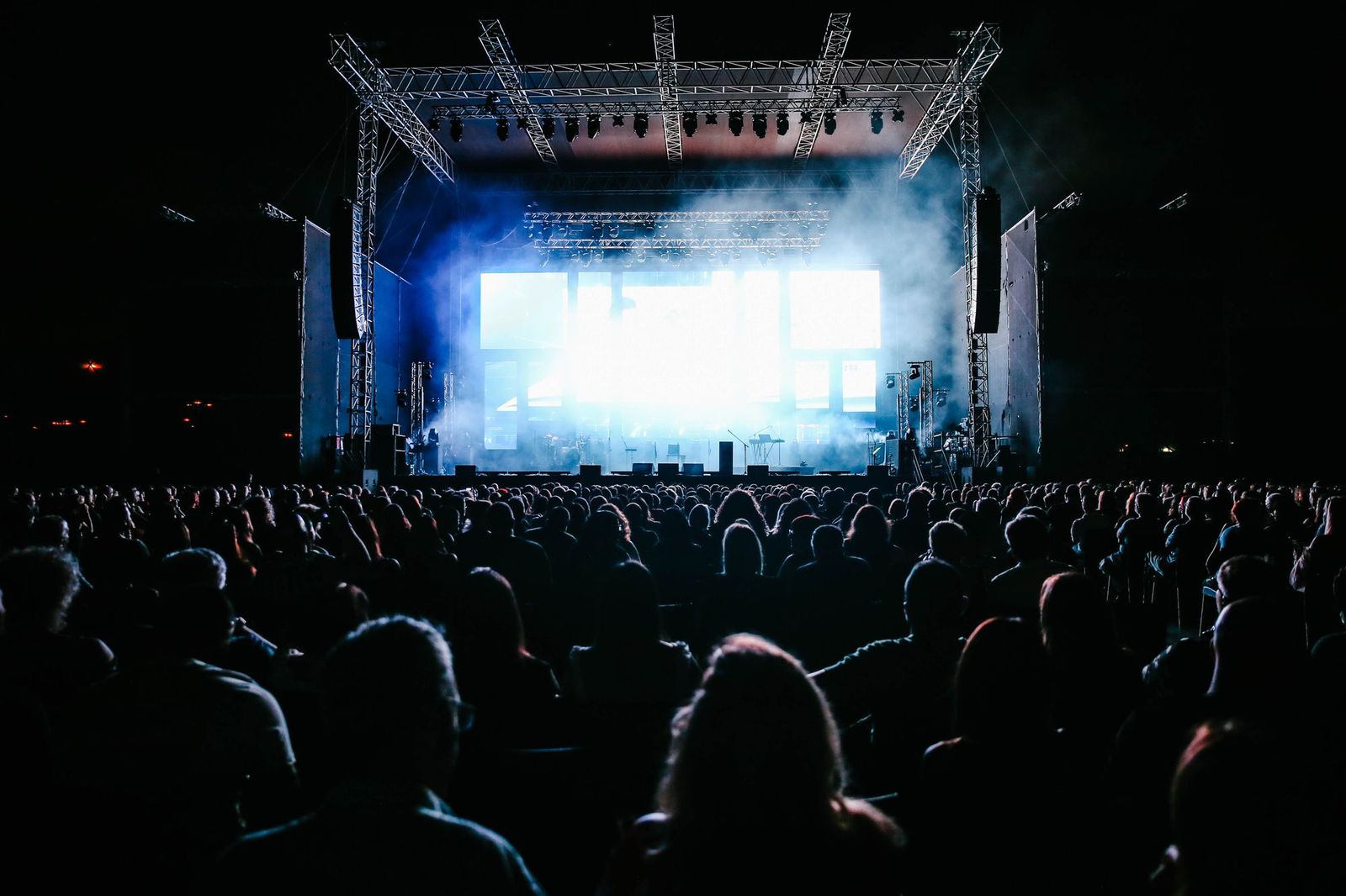 A packed outdoor crowd facing a brightly lit concert stage at night, haze glowing under the stage lights